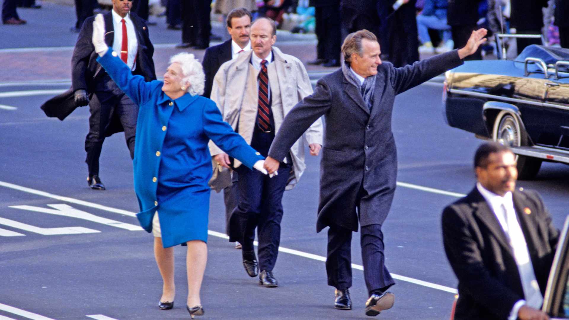 First Lady Barbara Bush (wearing a bright blue coat) and US President George HW Bush (in a gray overcoat and scarf) hold hands, smile and wave as they lead the Inaugural Parade along Pennsylvania Avenue, Washington DC, January 20, 1989. In front of and behind them in the middle of the street are various Secret Service agents.