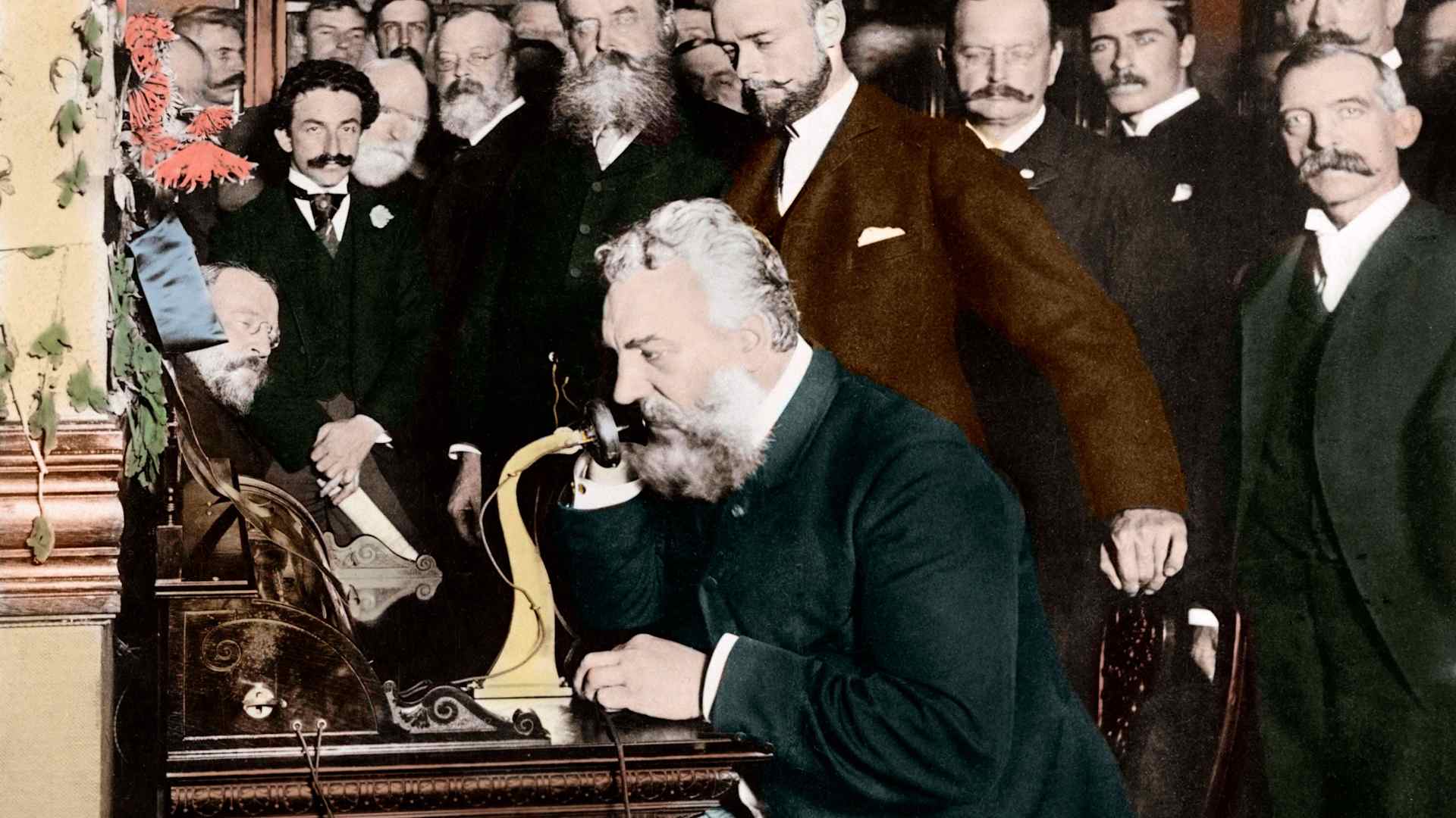 A group of businessmen watch inventor Alexander Graham Bell as he opens the New York-Chicago telephone line.
