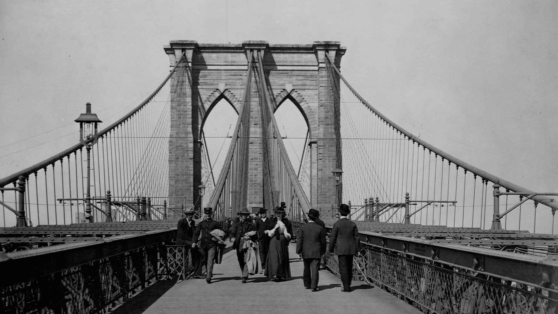 Men and women stroll along the promenade deck of the Brooklyn Bridge.