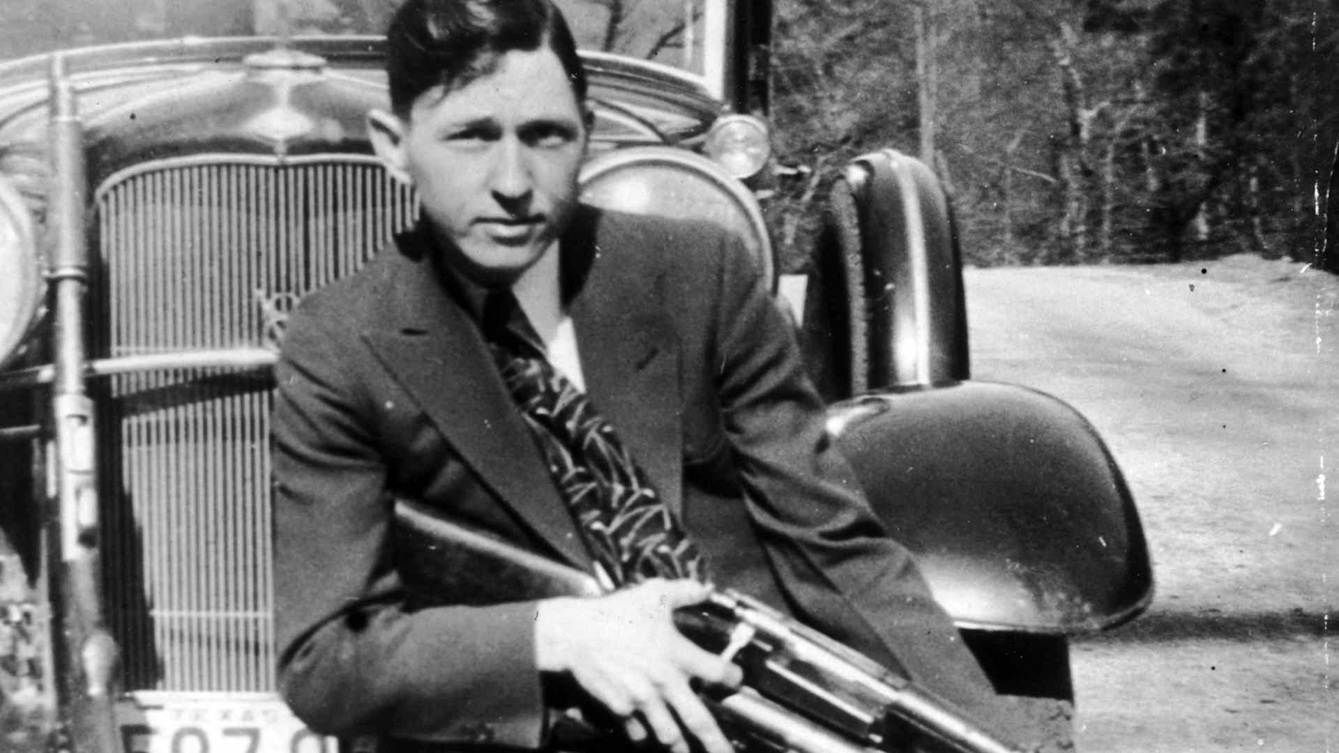 Clyde Barrow, holding a firearm while sitting on the front fender of a car circa 1933.