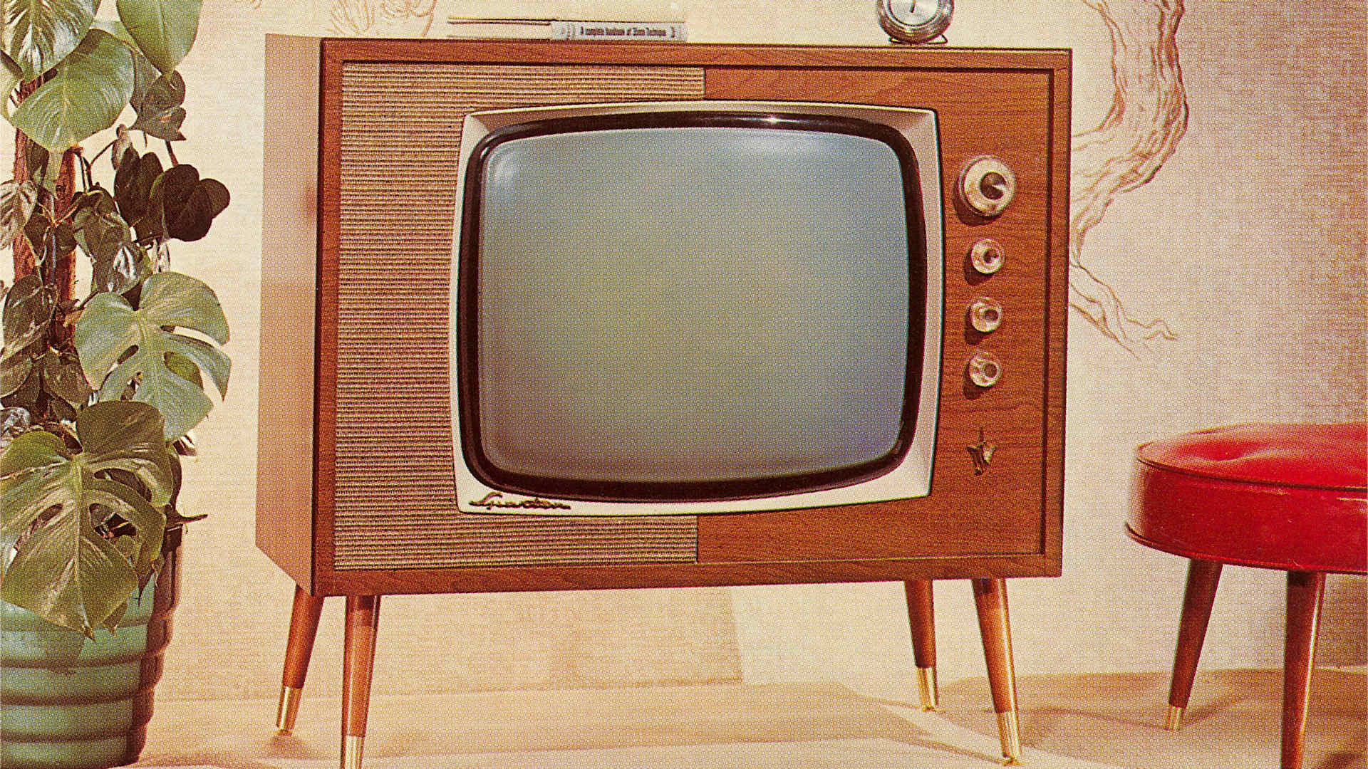 A 1960s-era TV set on legs is shown between a potted plant and red footstool.