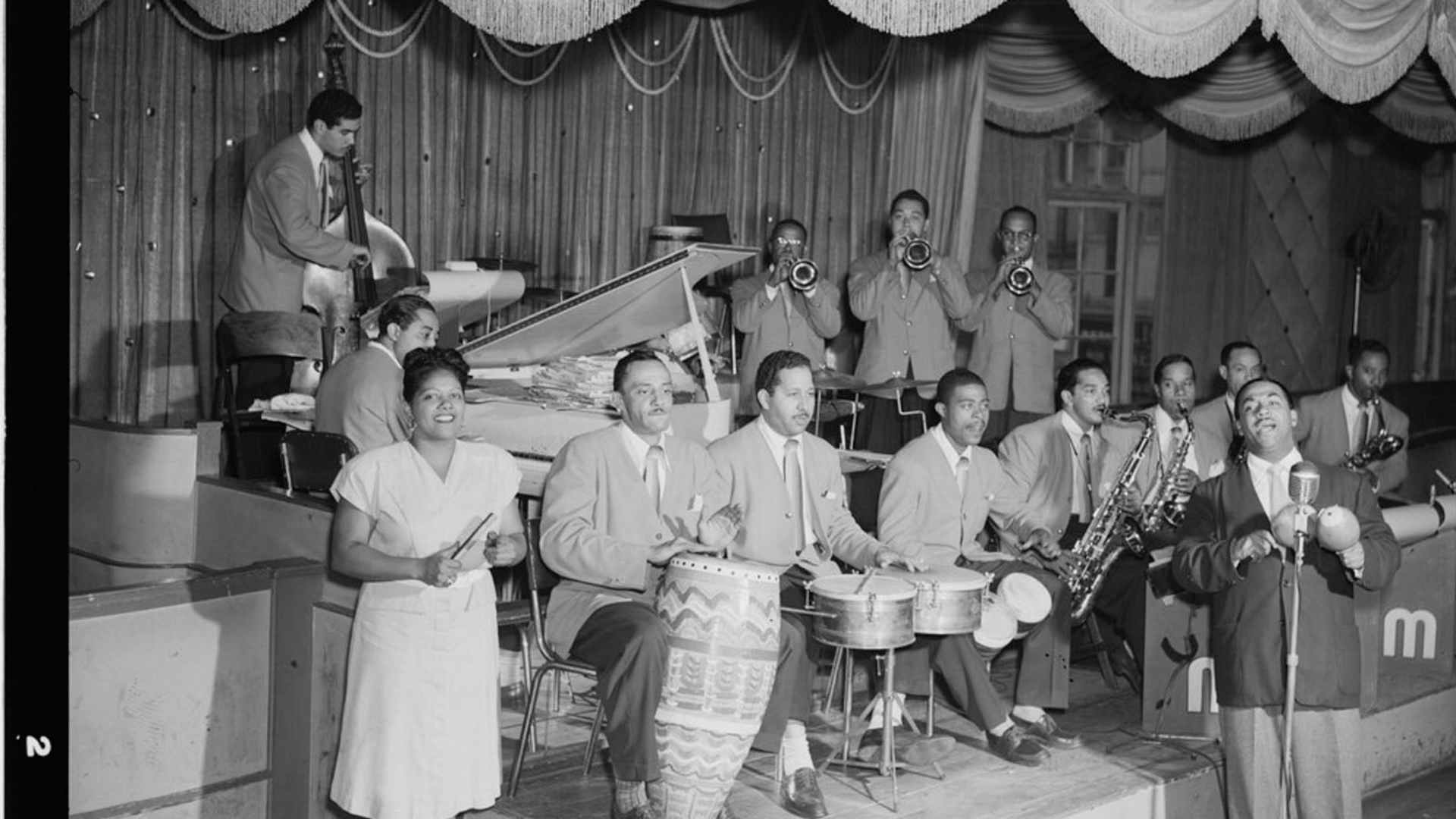 Portrait of Machito (at mic, right), with his Afro-Cuban mambo orchestra, including: José Mangual (bongos), Carlos Vidal Bolado (conga), Mario Bauzá (far left trumpeter), Ubaldo Nieto (timbales) and Graciella Grillo (front left), at the Glen Island Casino, New Rochelle, New York, c. July 1947