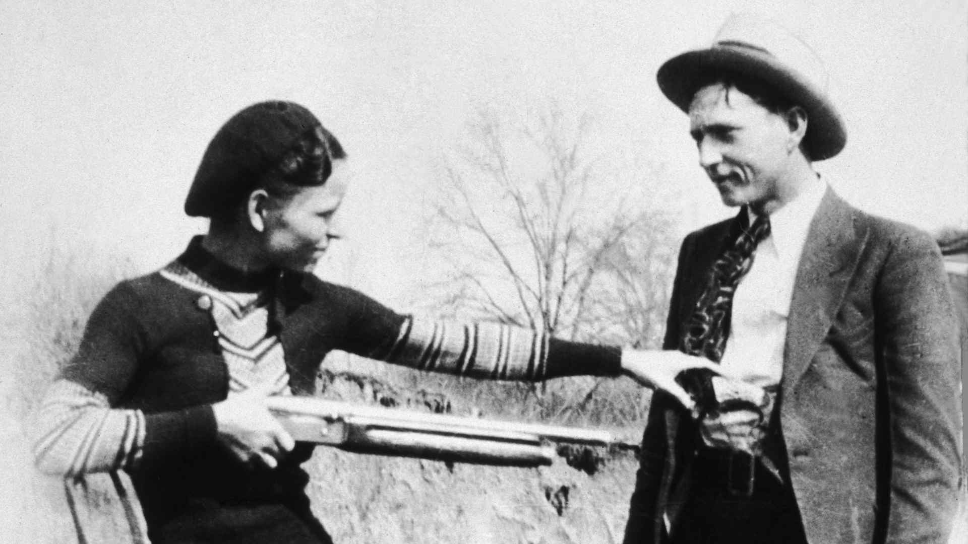 circa 1932: American criminal Bonnie Parker (1910 - 1934) aims a shotgun at her partner, Clyde Barrow (1909 - 1934) while clowning beside an automobile.