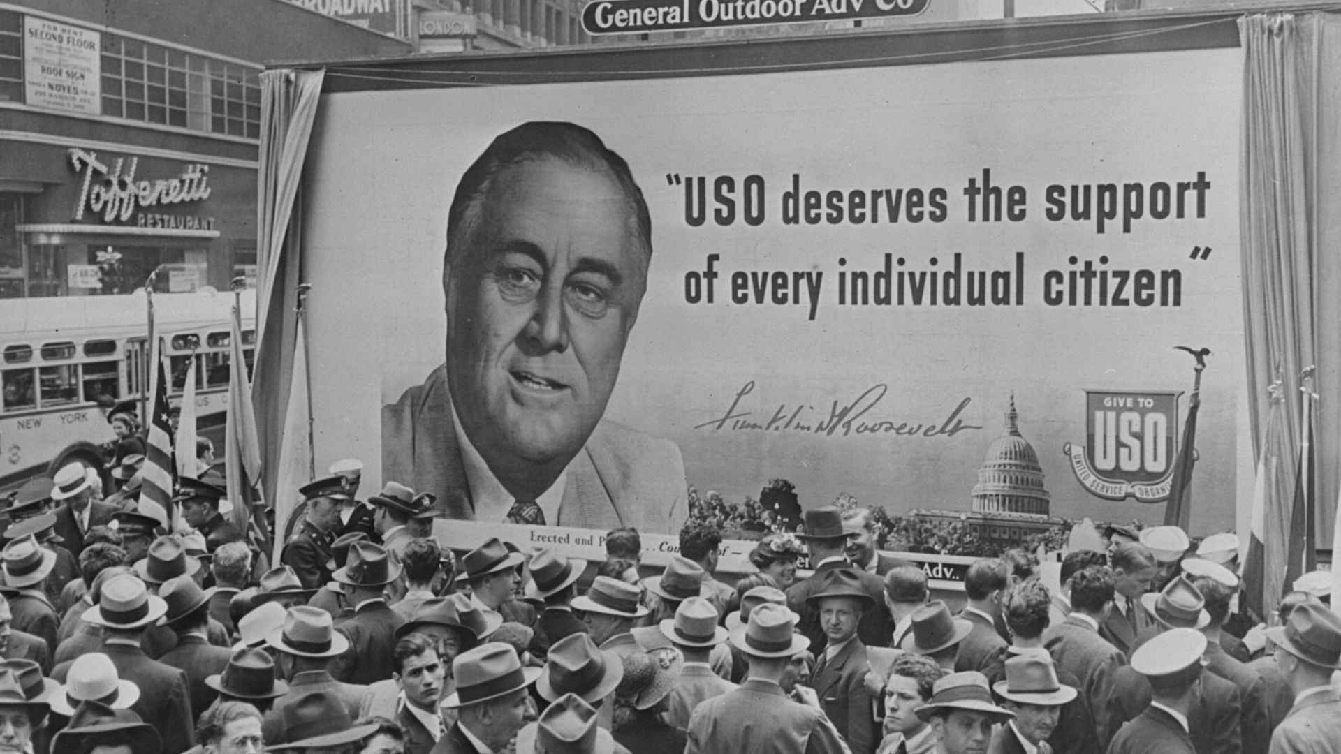 circa 1941: A crowd gathered in front of a poster for an armaments drive featuring a portrait of American President Franklin Delano Roosevelt.