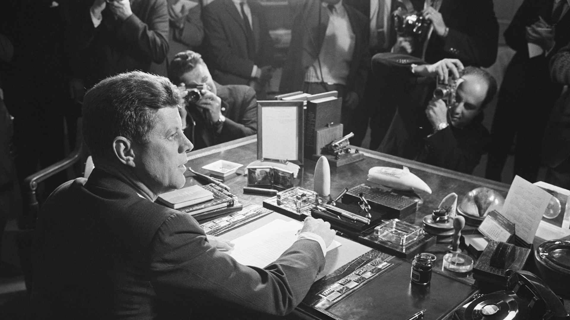 Reporters take pictures of President Kennedy behind his desk, after signing the arms embargo against Cuba. The embargo effectively quarantined Cuba.