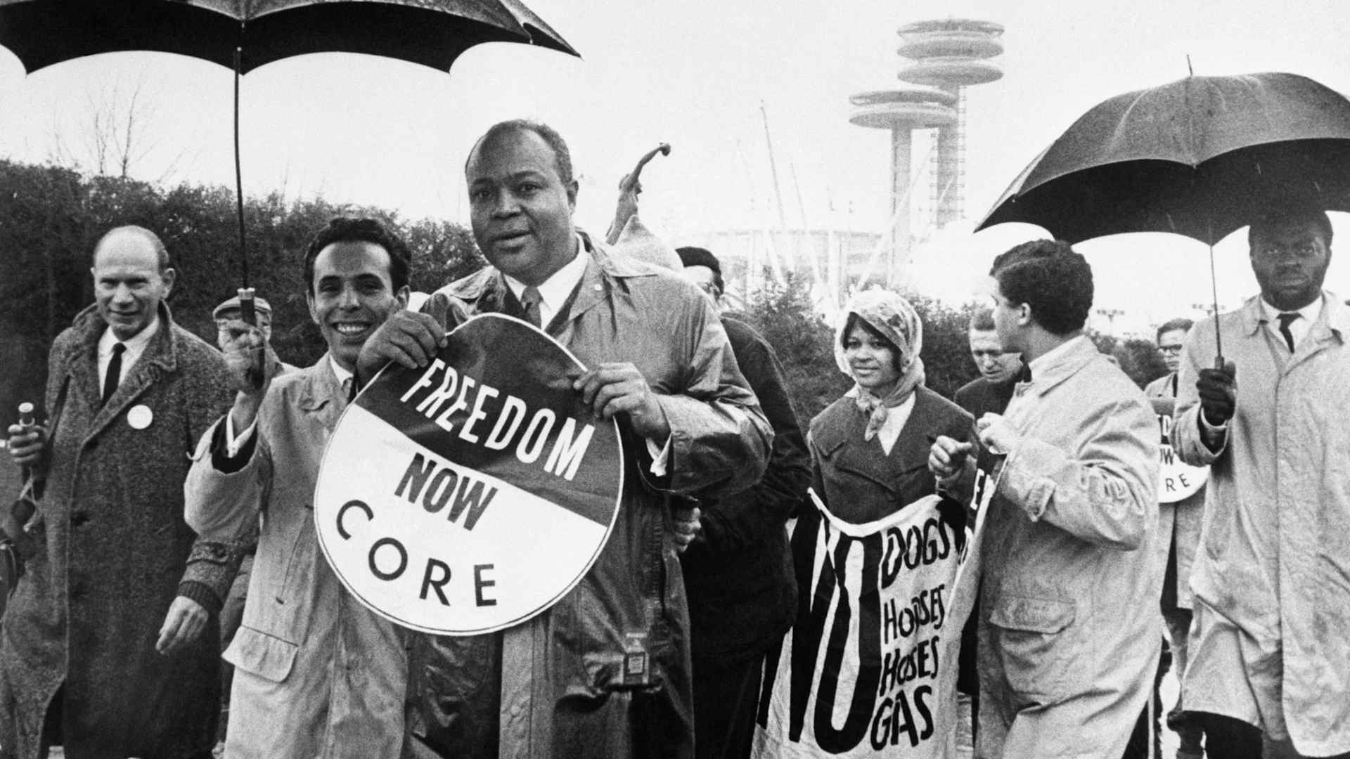 James Farmer, national director of the Congress of Racial Equality, holds a large sign as he leads a CORE demonstration during opening day ceremonies at the World's Fair. In the background is the New York State Pavilion.