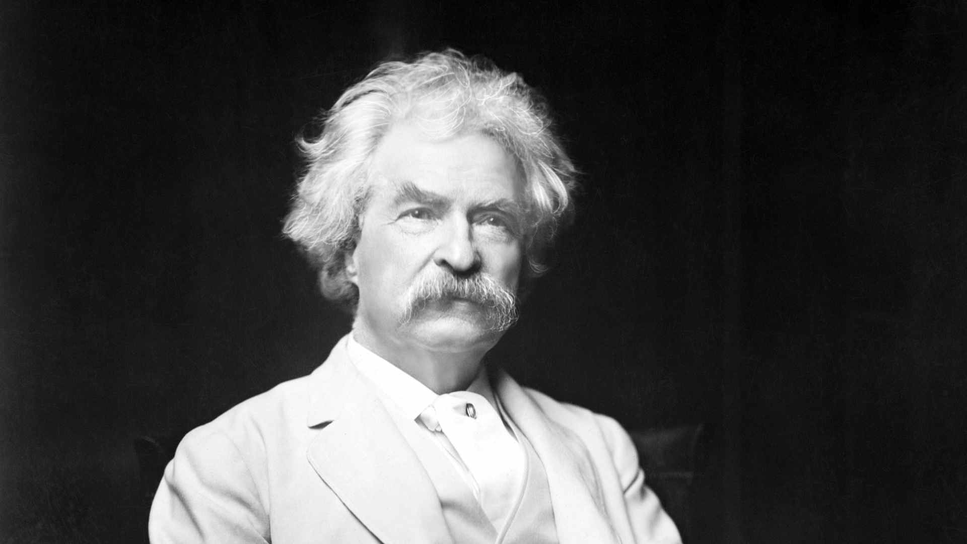 Mark Twain (Samuel Clemens) 1885-1910, lecturer and author of great literature. In this 3/4 length photograph he's seated holding a cigar, in a white suit. Undated.