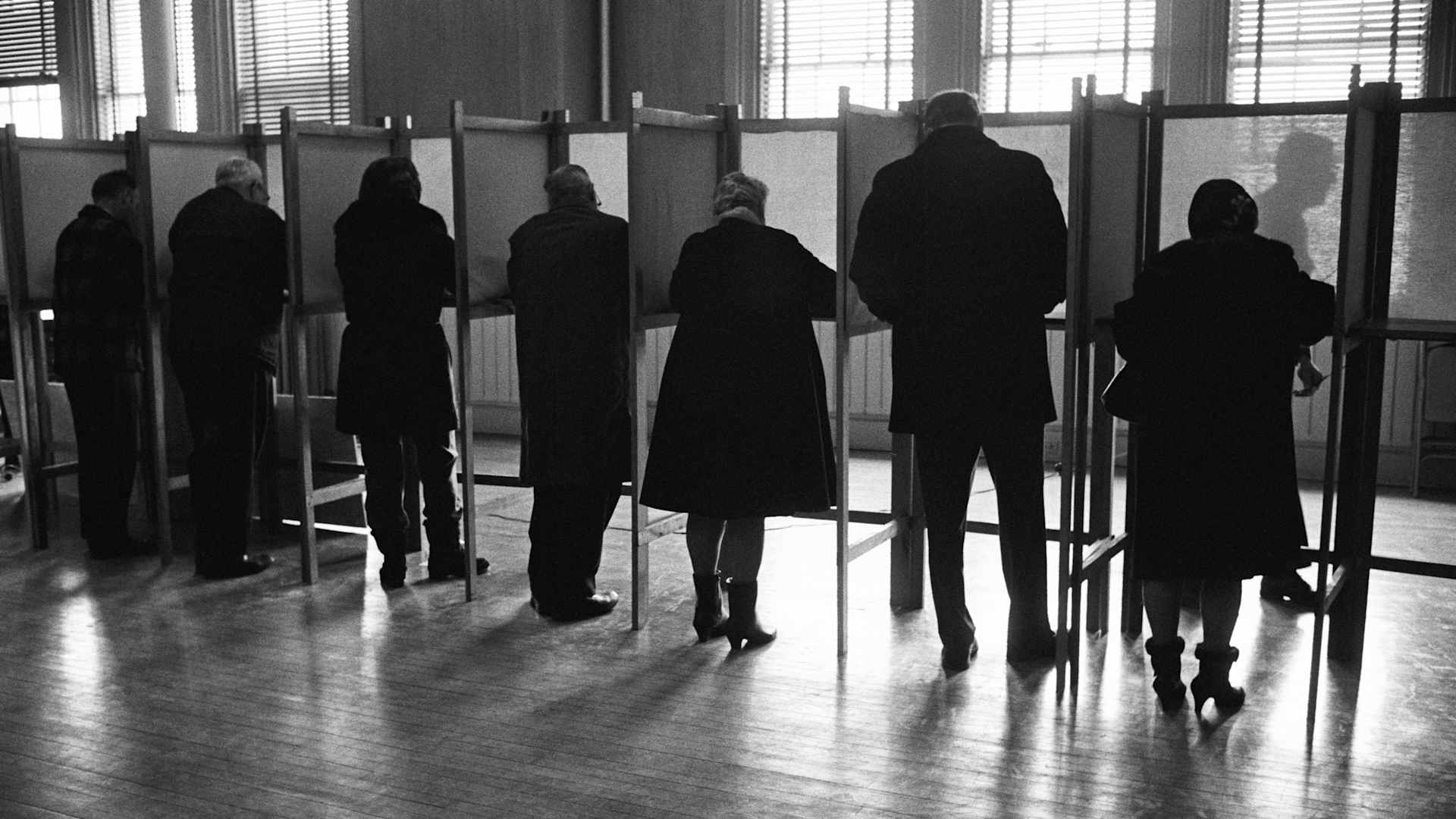 Circa 1970s: Men and women standing side by side in private voting booths, filling out election ballot papers.