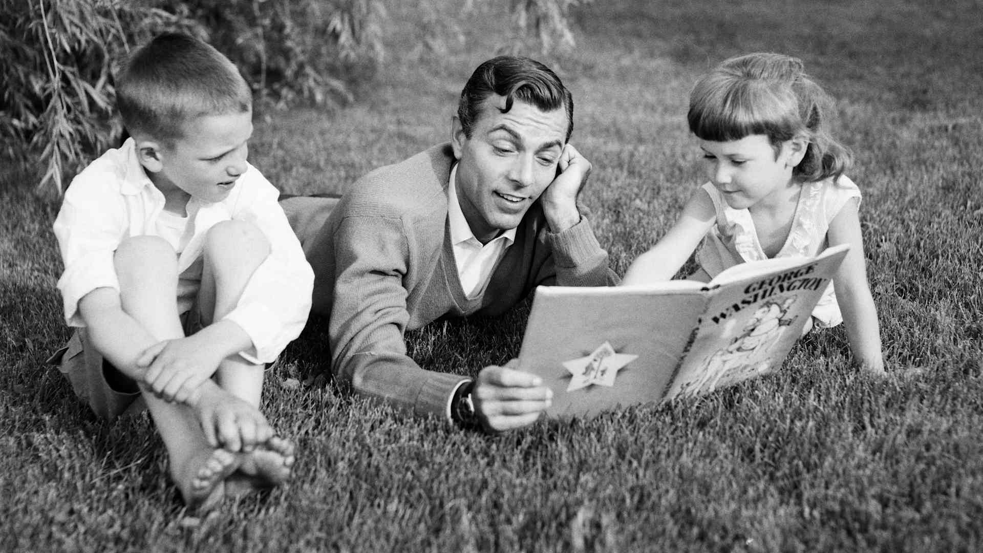 1950s father lying in the grass reading to his son and daughter