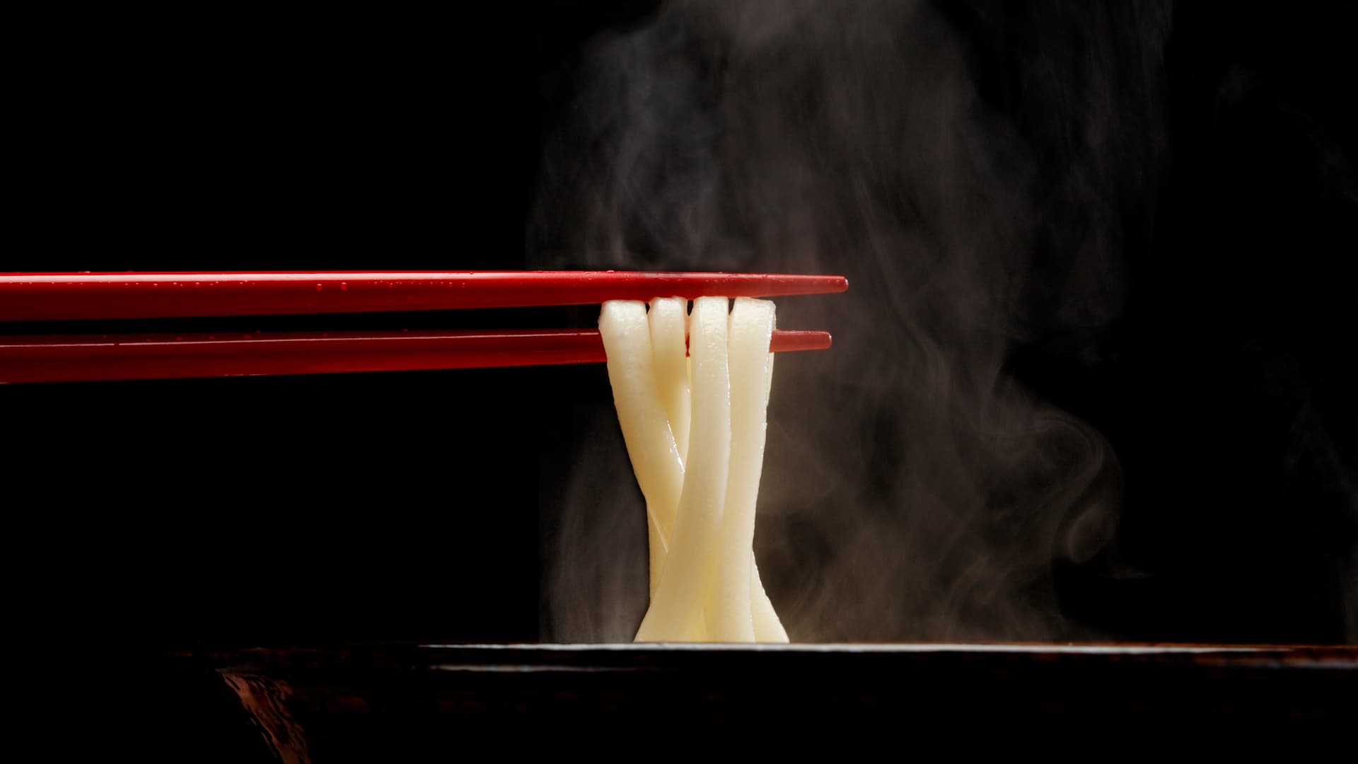 Chopsticks picking up noodles from a steaming bowl.