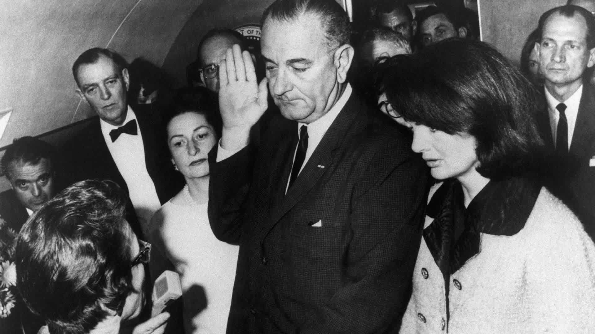 Vice President Lyndon B. Johnson is sworn in to the office of the Presidency aboard Air Force One in Dallas, Texas, hours after the assassination of President John F. Kennedy. Johnson is flanked by wife, Lady Bird Johnson (L), and First Lady Jacqueline Kennedy during the ceremony, which is being administered by U.S. District Judge Sarah Hughes. At farthest left in the background is Jack Valenti.