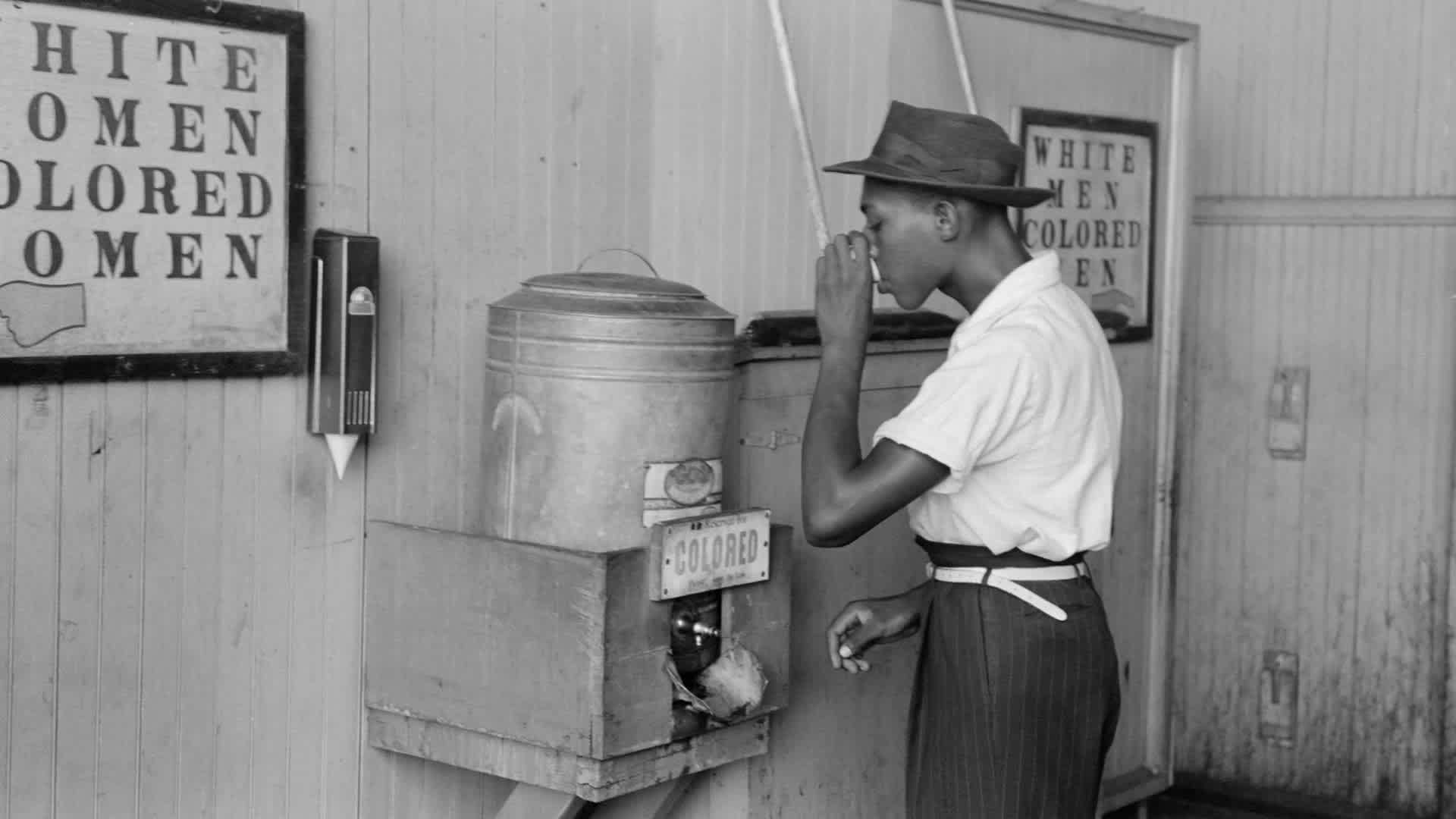 A man drinking water at a water cooler labeled 'Colored' in a bus terminal in Oklahoma City, Oklahoma, July 1939.