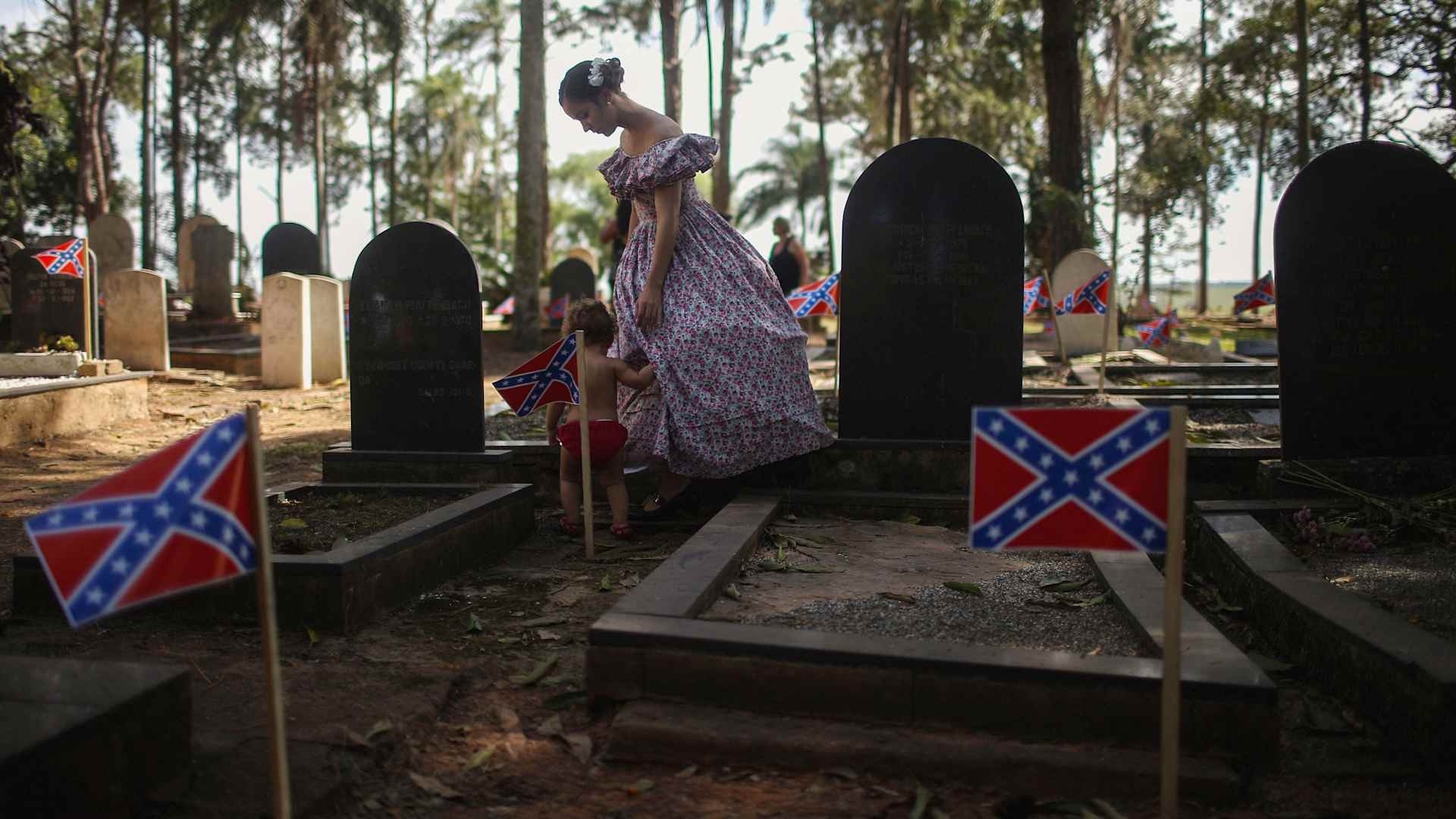 A woman dressed in traditional hoop skirt walks past graves marked with Confederate flags in the American Cemetery during the annual Festa Confederada in Santa Barbara d'Oeste, Brazil, 2016. The festival is put on by Brazilian descendants of U.S. confederates who fled to Brazil during Reconstruction following the end of the Civil War.