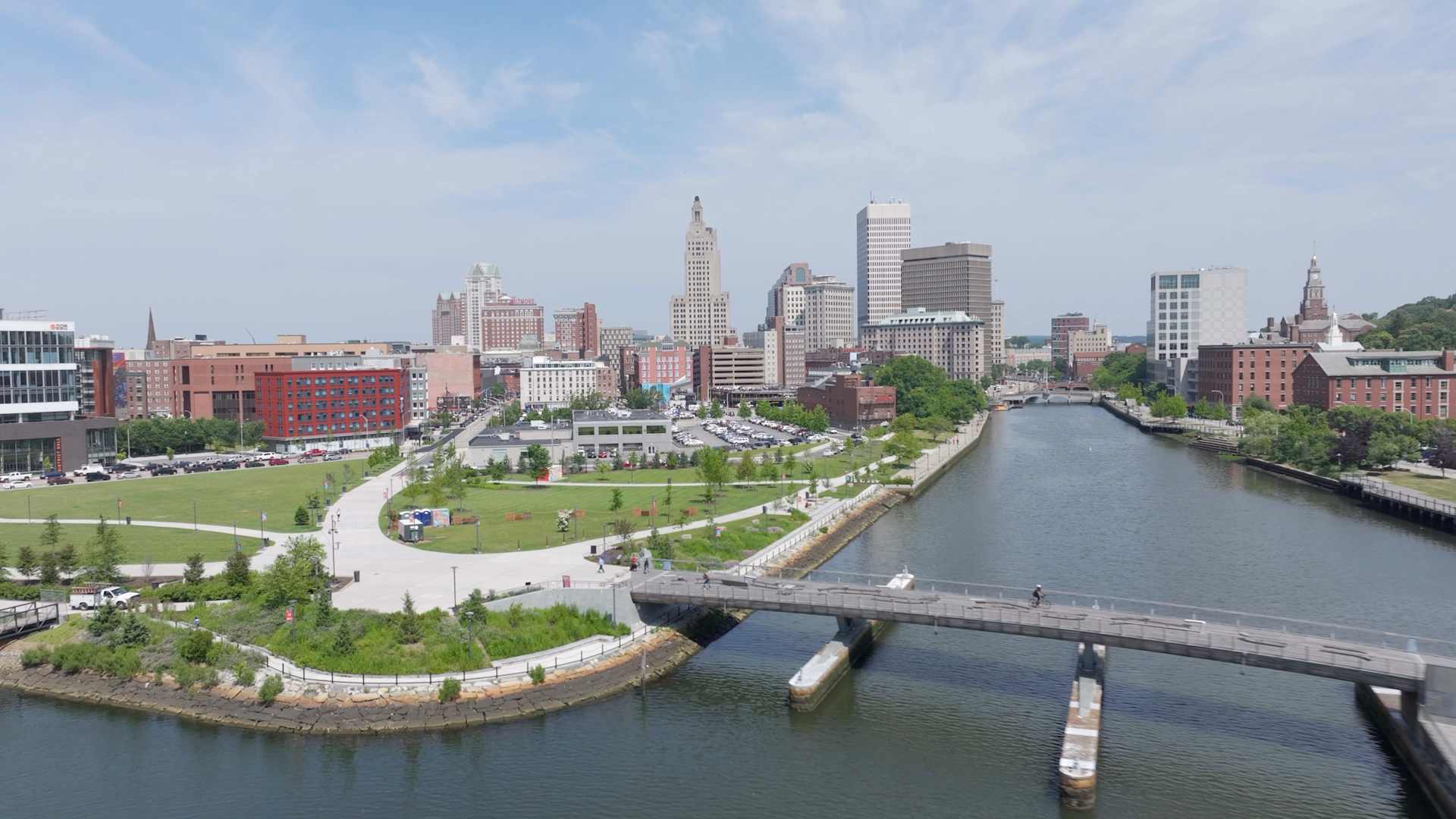 A cityscape with a canal in the foreground, surrounded by a park-like setting, and a skyline of tall buildings in the background.