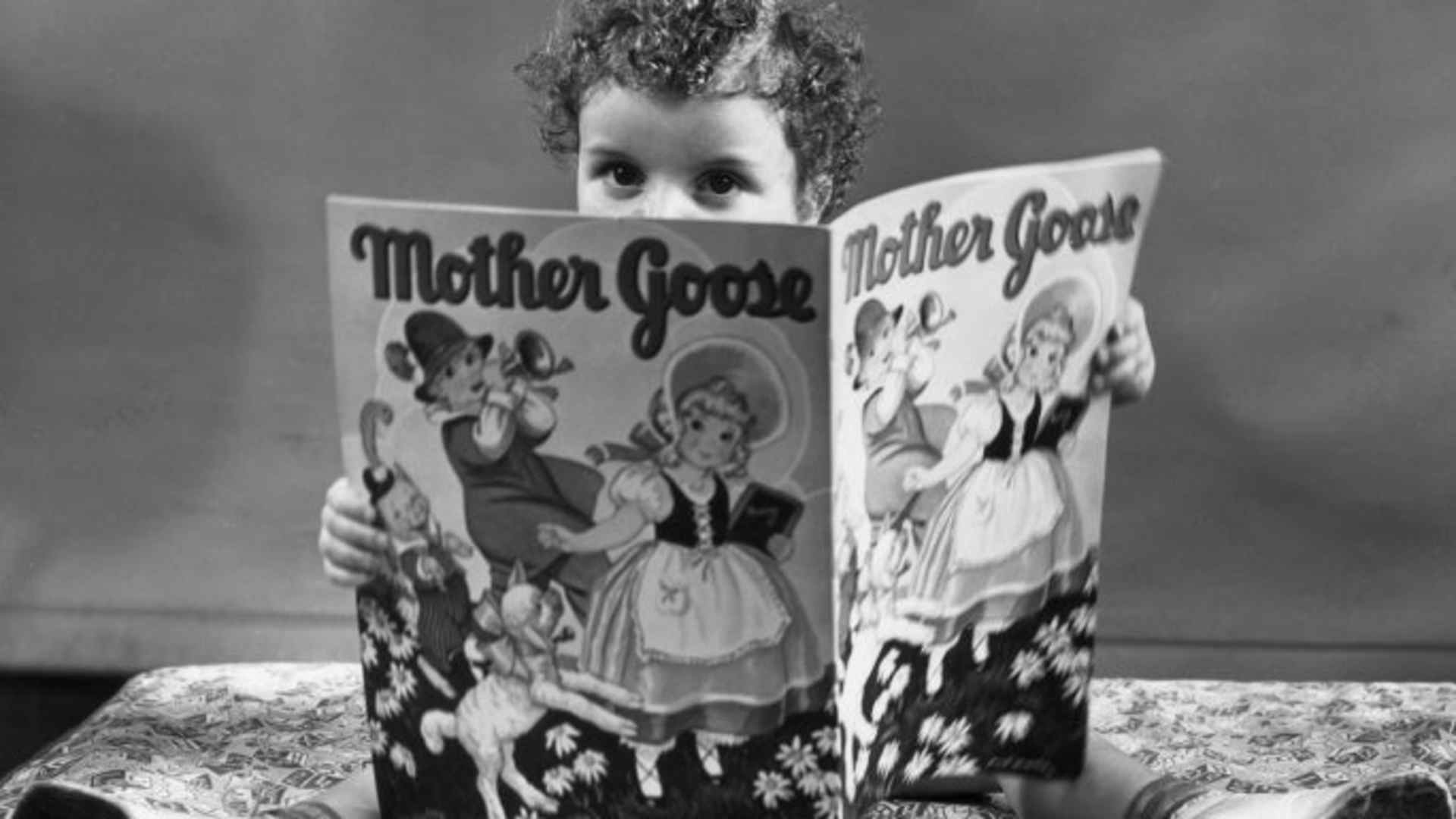 A young girl sits on top of her bed, reading a Mother Goose storybook.
