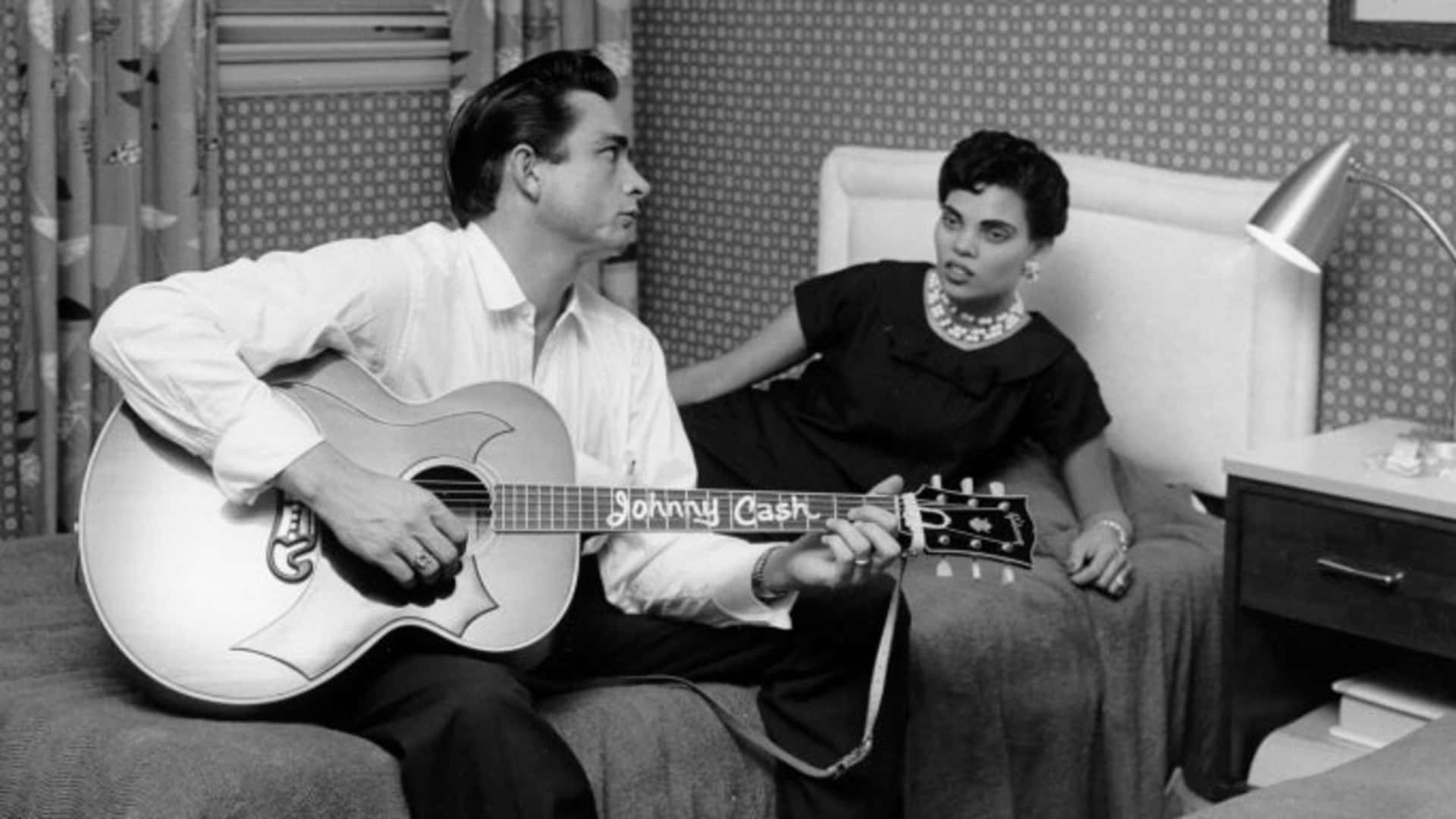 Country singer/songwriter Johnny Cash sits on a bed playing acoustic guitar to his first wife Vivian Liberto