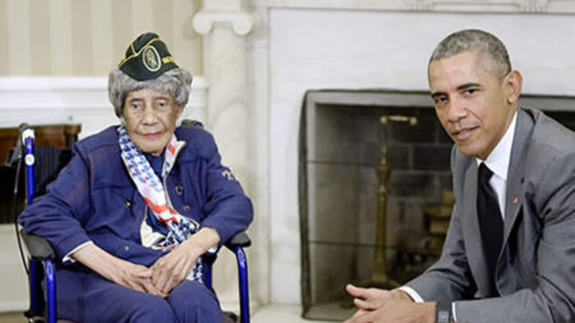 The image shows an elderly woman in a wheelchair wearing a military uniform and a man in a suit sitting next to her, both appearing to be engaged in conversation.