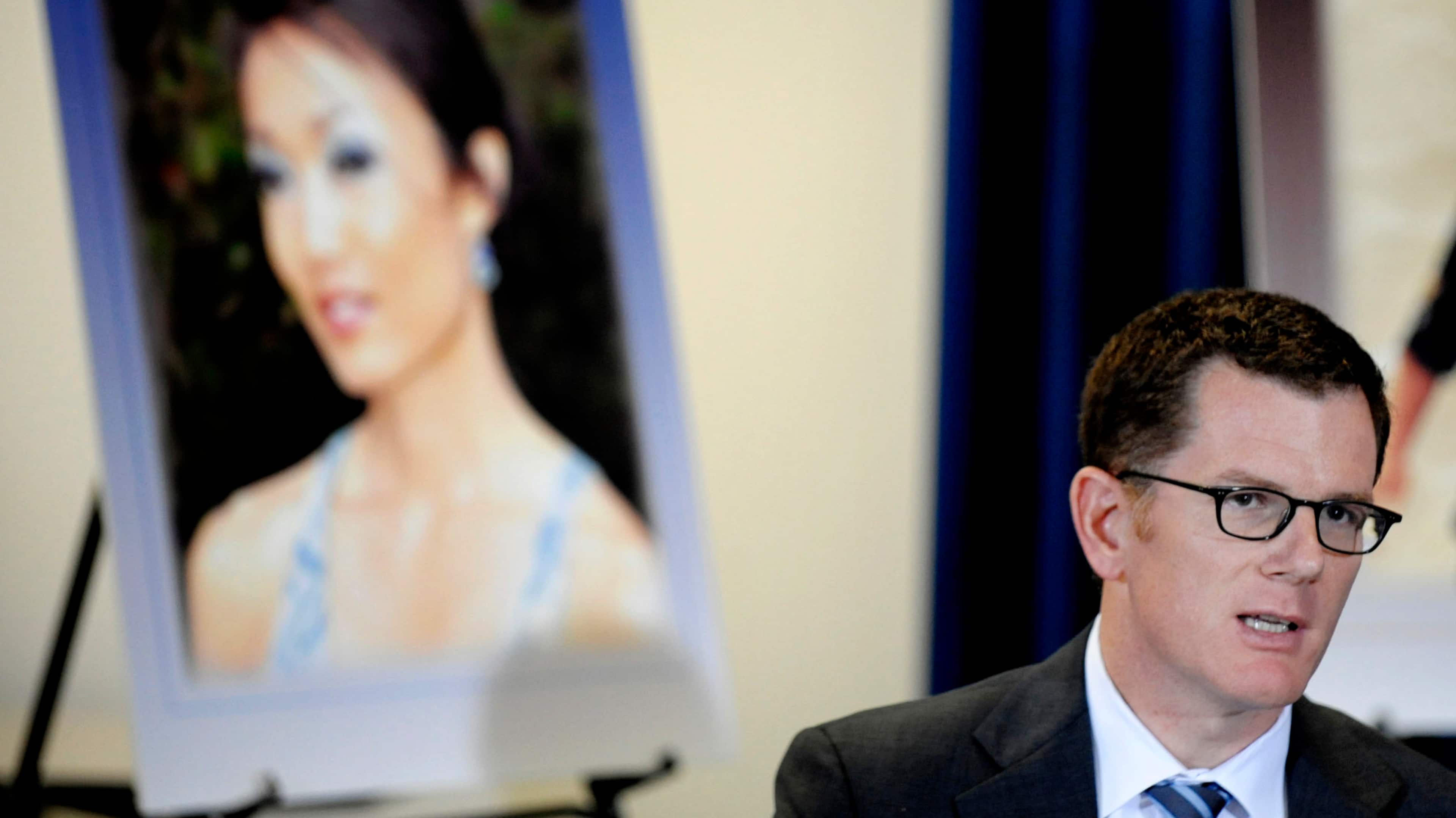 The image shows a man in a suit speaking in front of a large portrait of a woman.