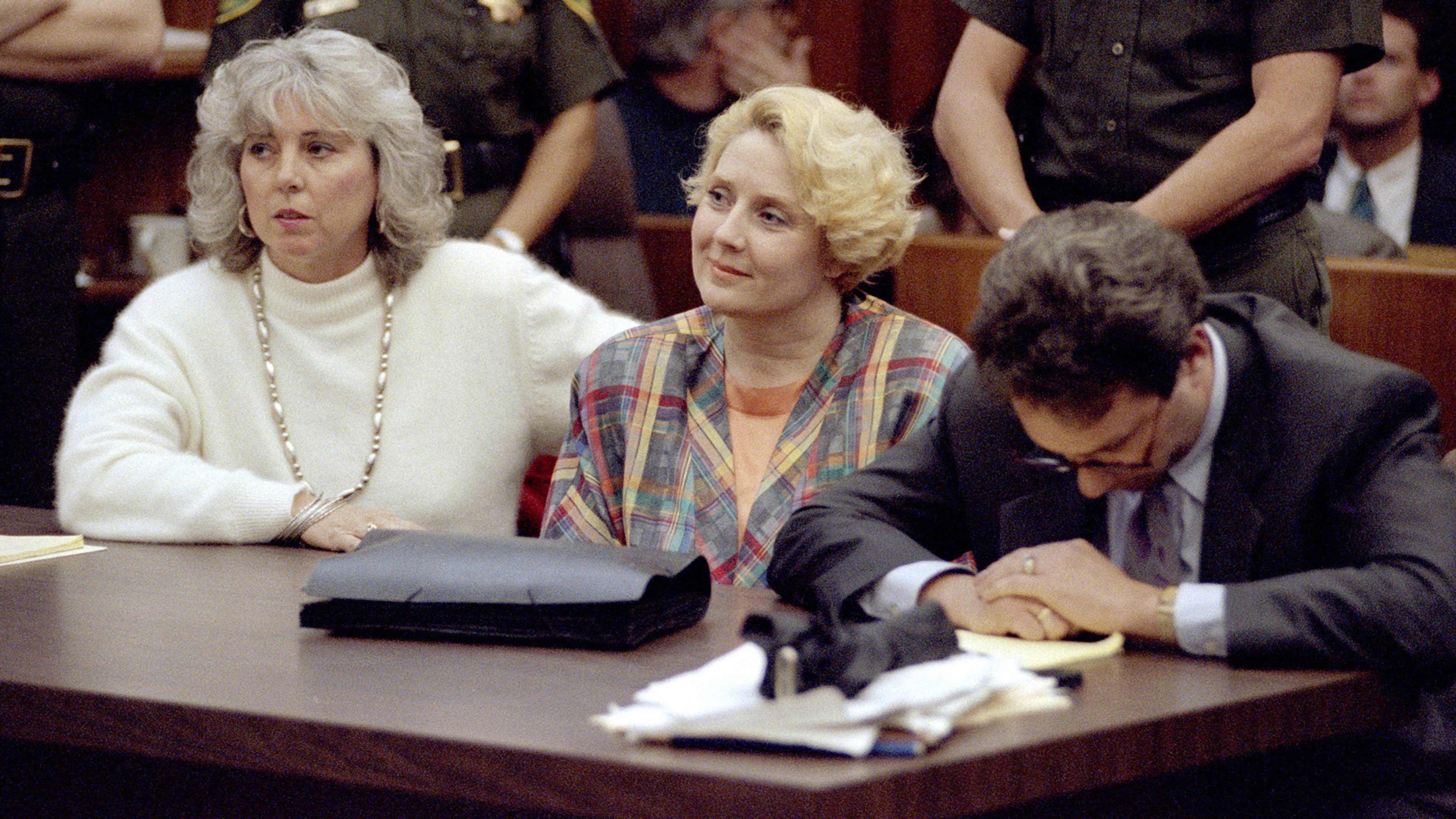 The image shows three people sitting at a table, with two women on the left and a man on the right. The women appear to be in formal attire, while the man is wearing a suit. The background suggests a formal setting, such as a courtroom or government office.