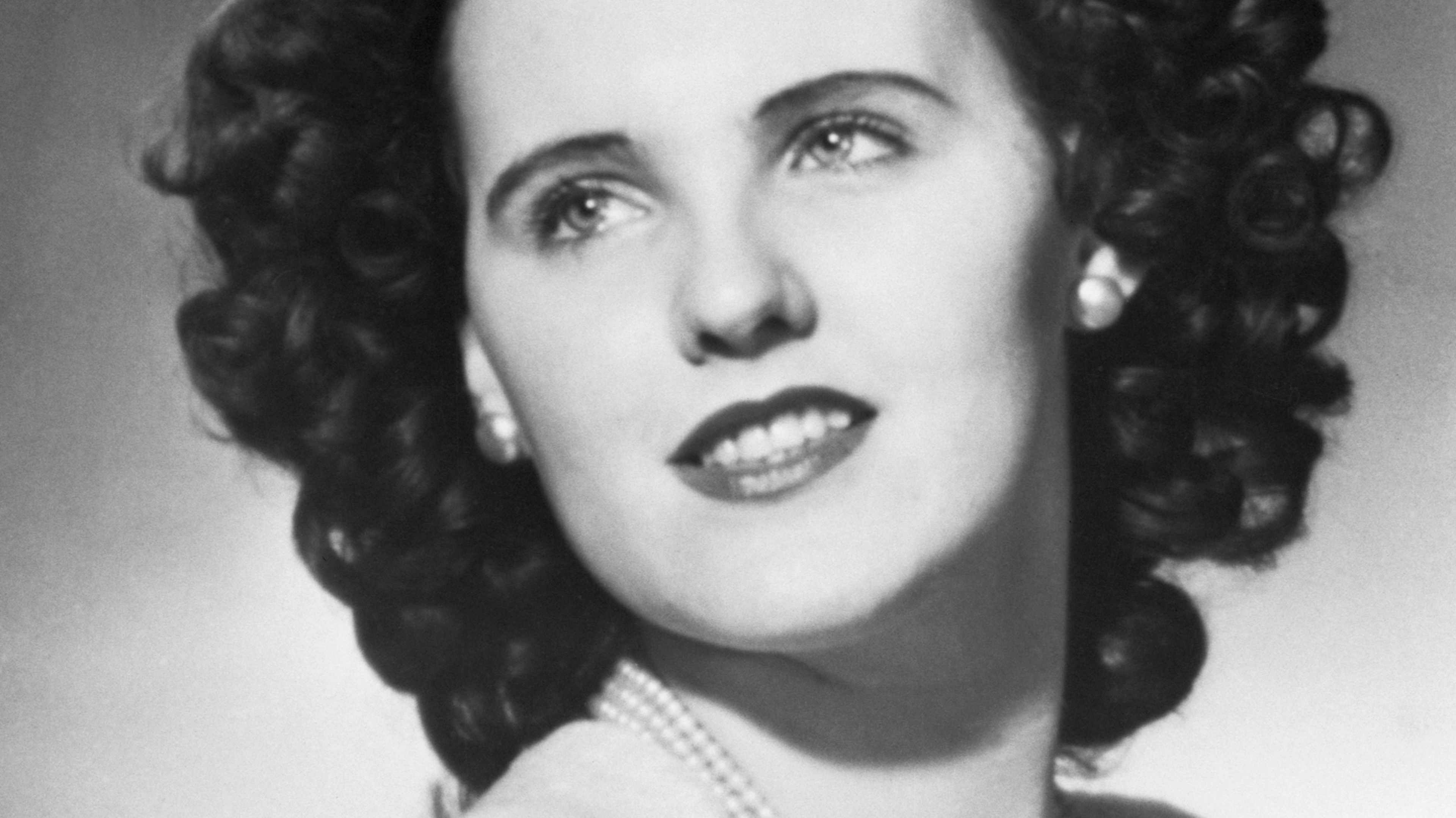 A black and white portrait of a woman with curly hair and an expressive facial expression, set against a plain background.
