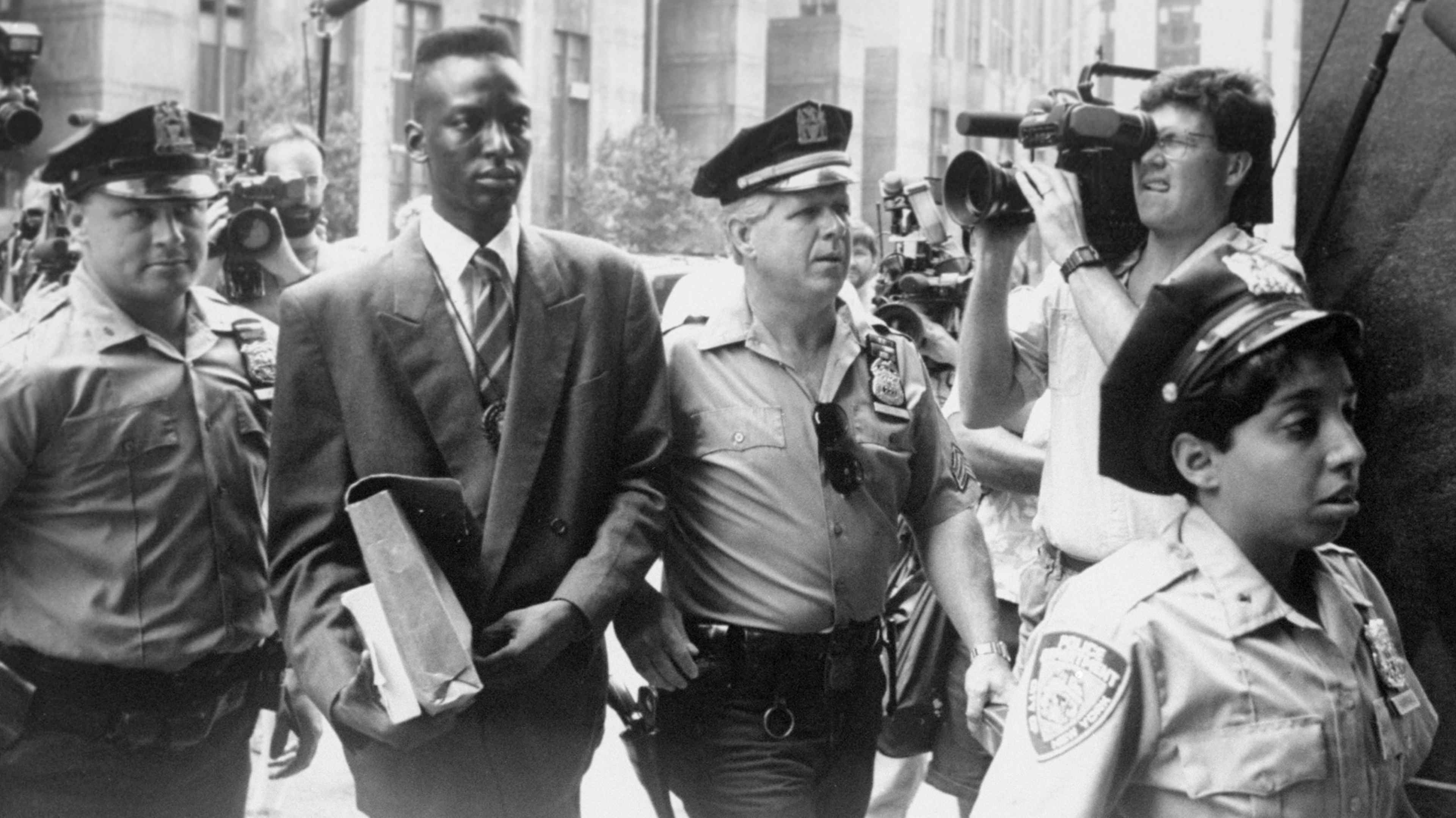 A black and white photograph depicting a group of men, some in uniform, walking together down a crowded city street, with onlookers and photographers visible in the background.
