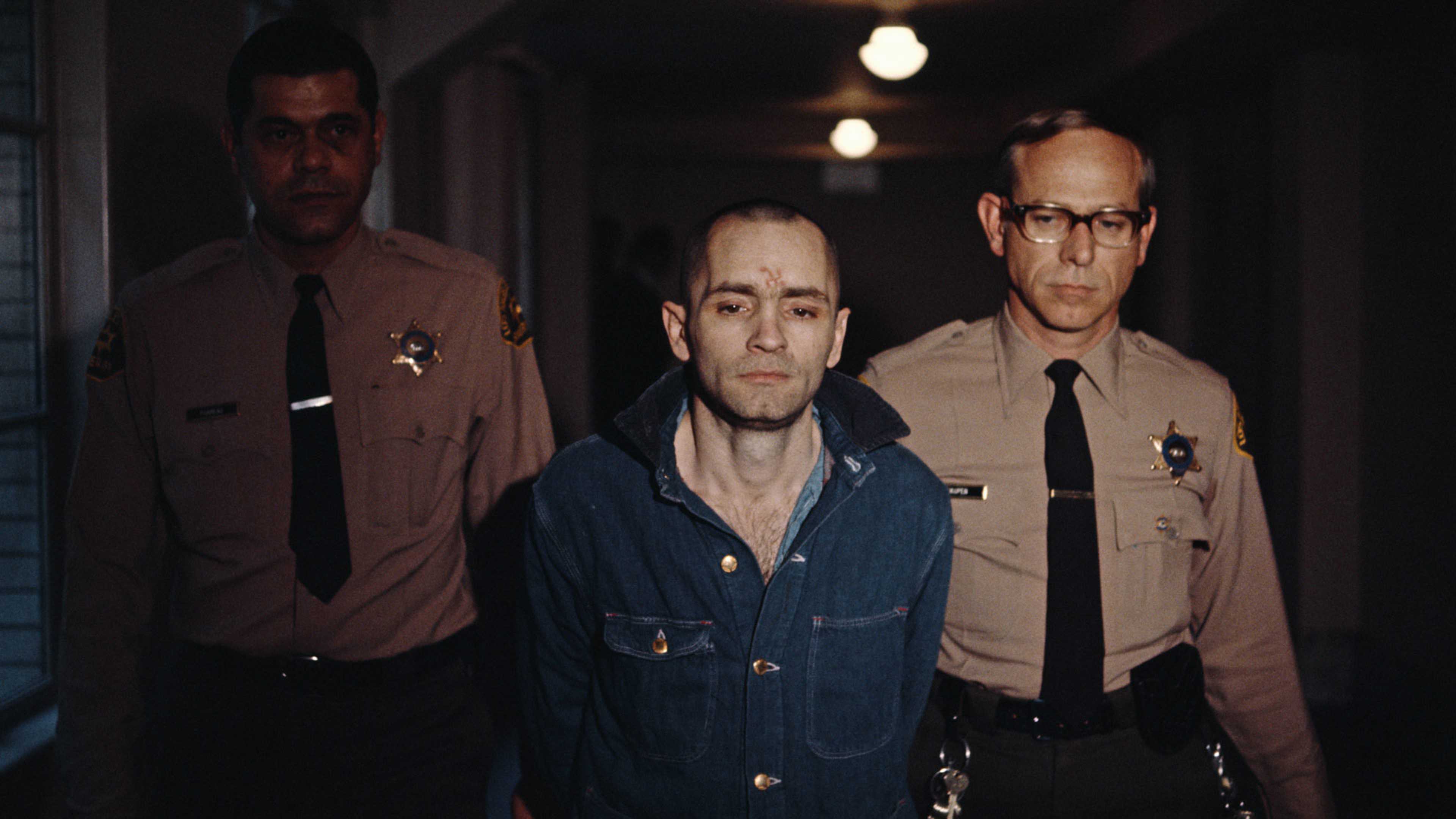 The image depicts three individuals, one in a blue jacket and two in uniform, walking together in a dimly lit setting.