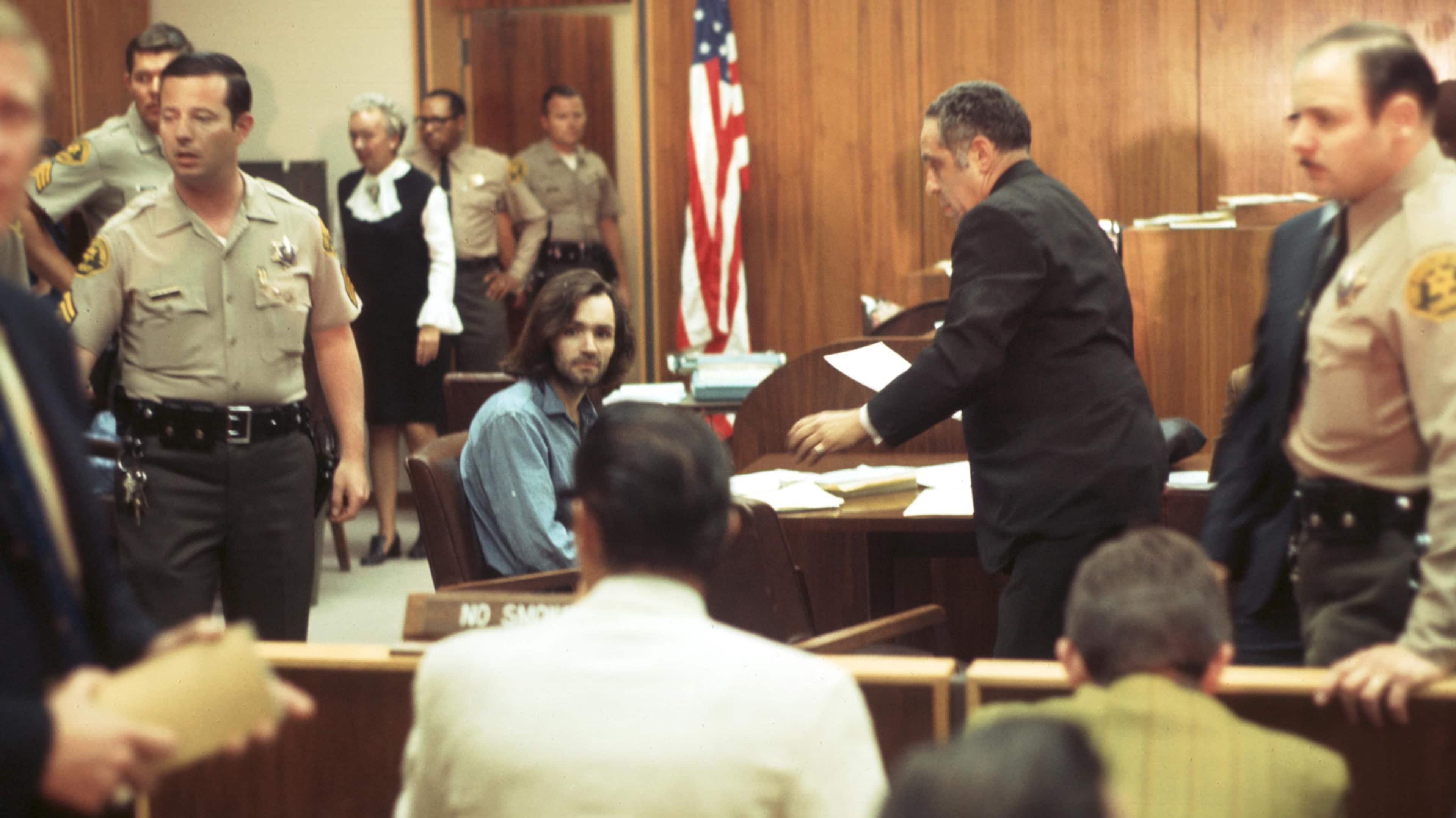 The image depicts a courtroom setting, with a judge presiding over a trial. Several individuals, including law enforcement officers and what appears to be a defendant, are present in the room.