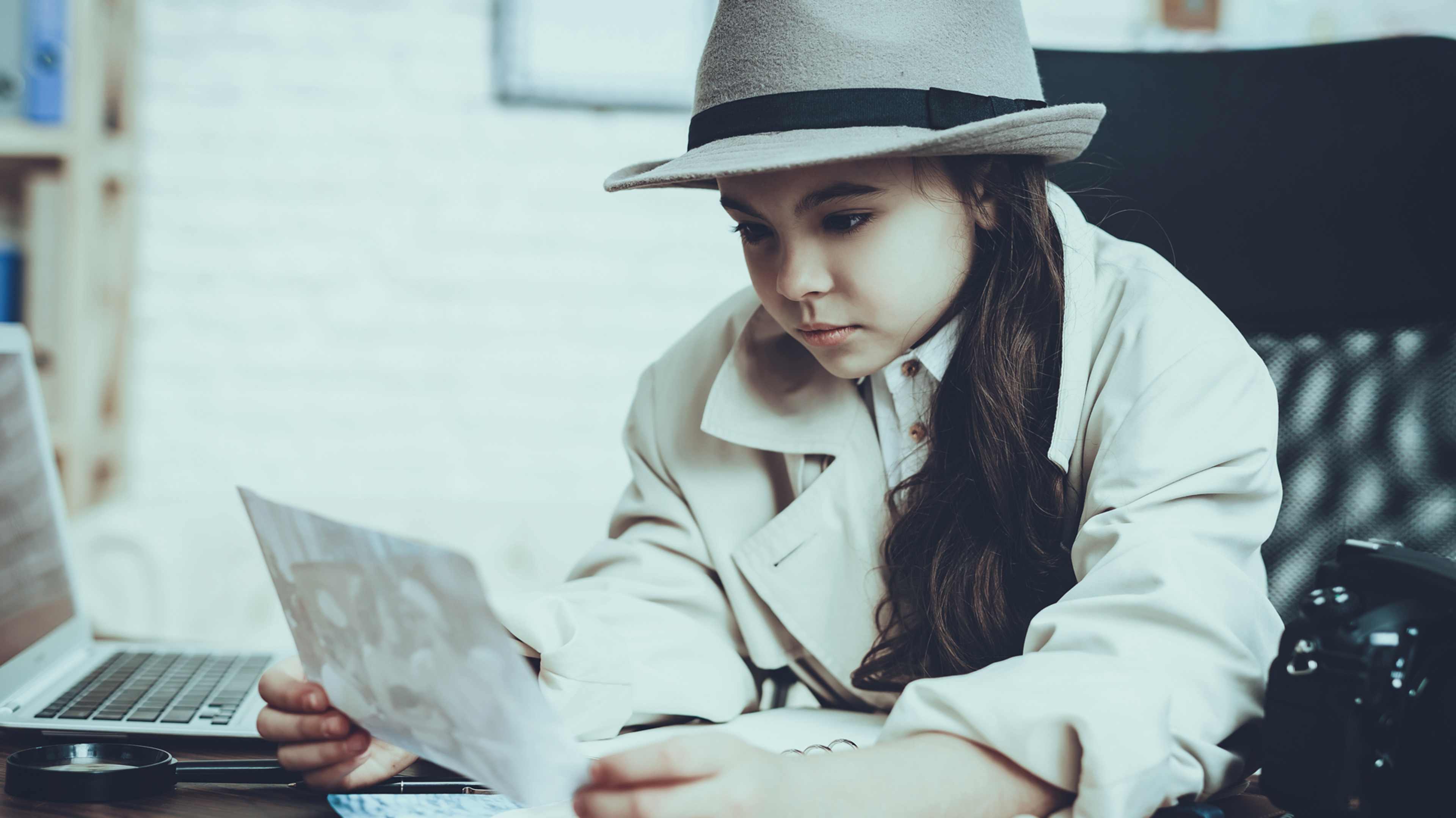 A young woman wearing a white coat and a wide-brimmed hat is sitting on a bench, intently focused on a laptop computer in front of her.