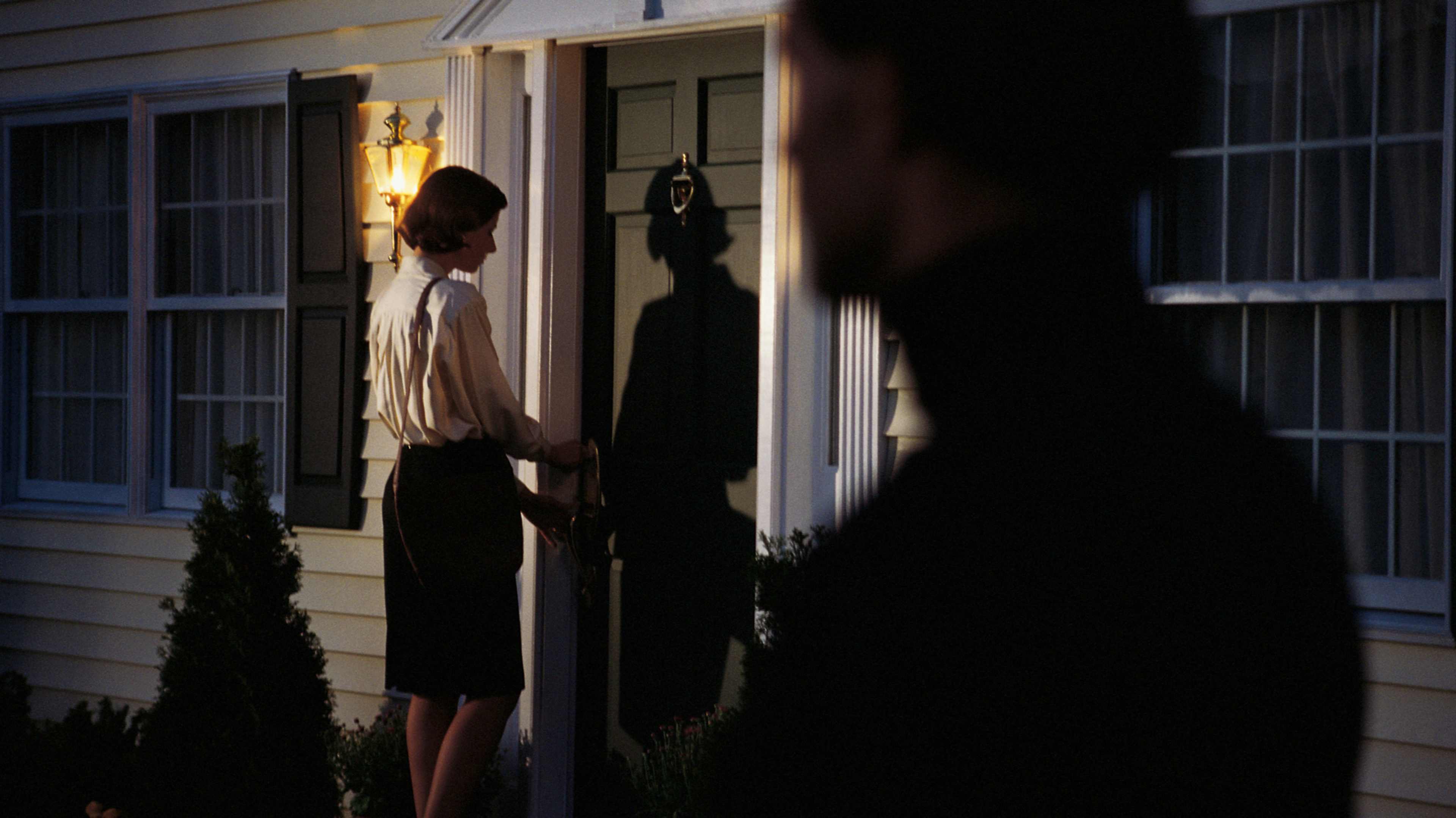 Two figures stand in the doorway of a dimly lit building, their silhouettes casting long shadows against the backdrop of the night.