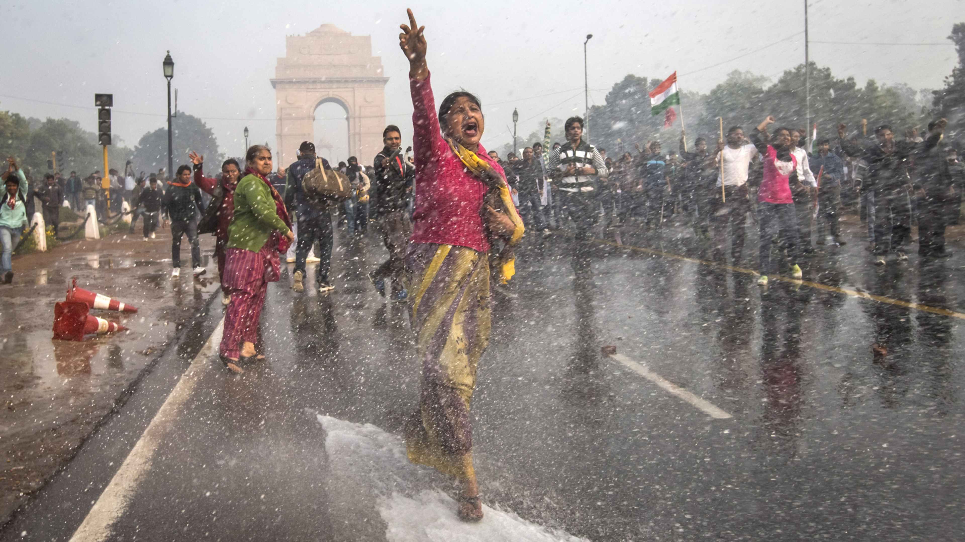 A group of people, dressed in colorful traditional attire, are walking through a rainy street in what appears to be a city, with the iconic India Gate visible in the background.