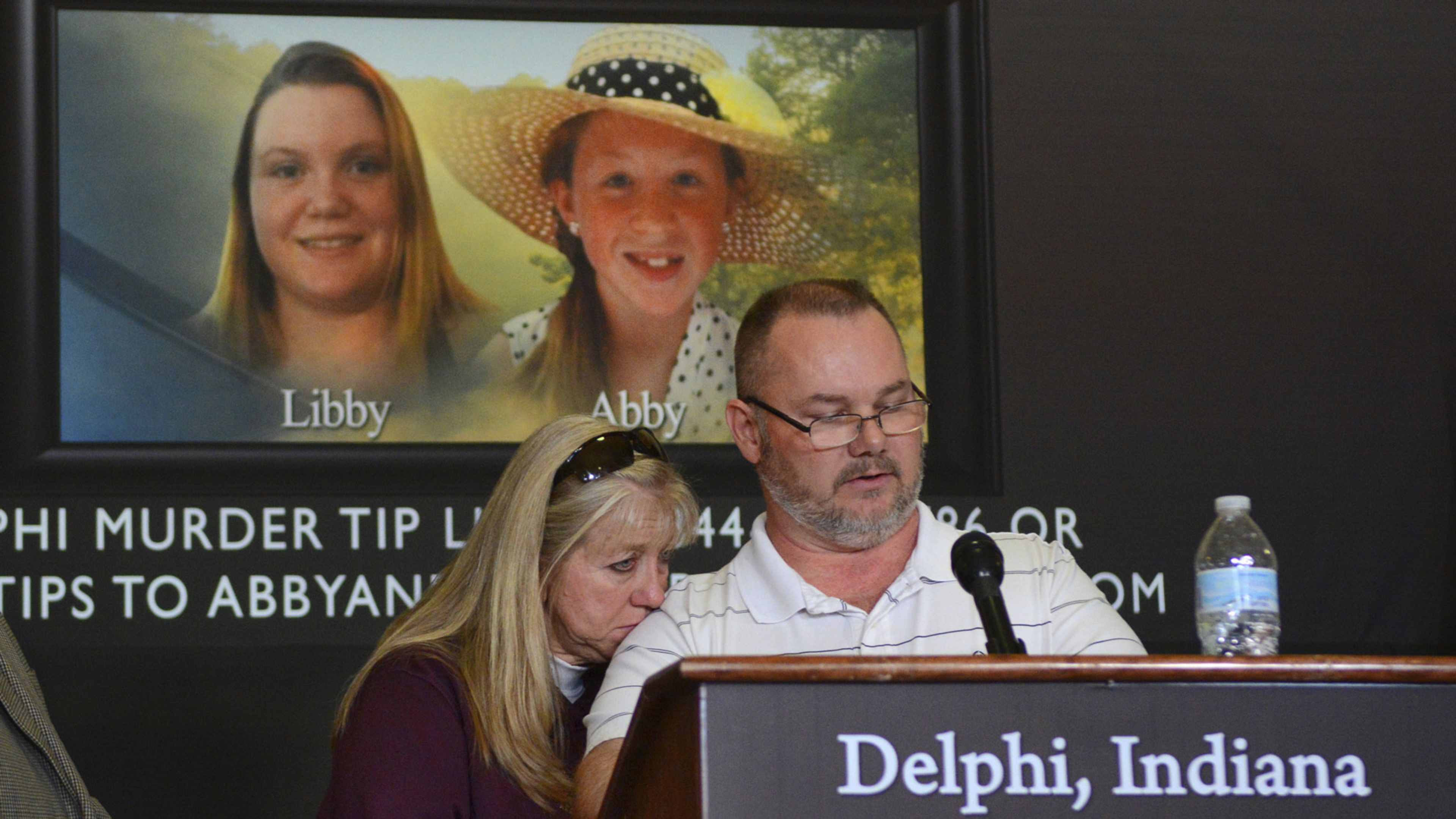 The image shows a man and a woman speaking at a podium, with a large screen behind them displaying a photograph of two smiling women.