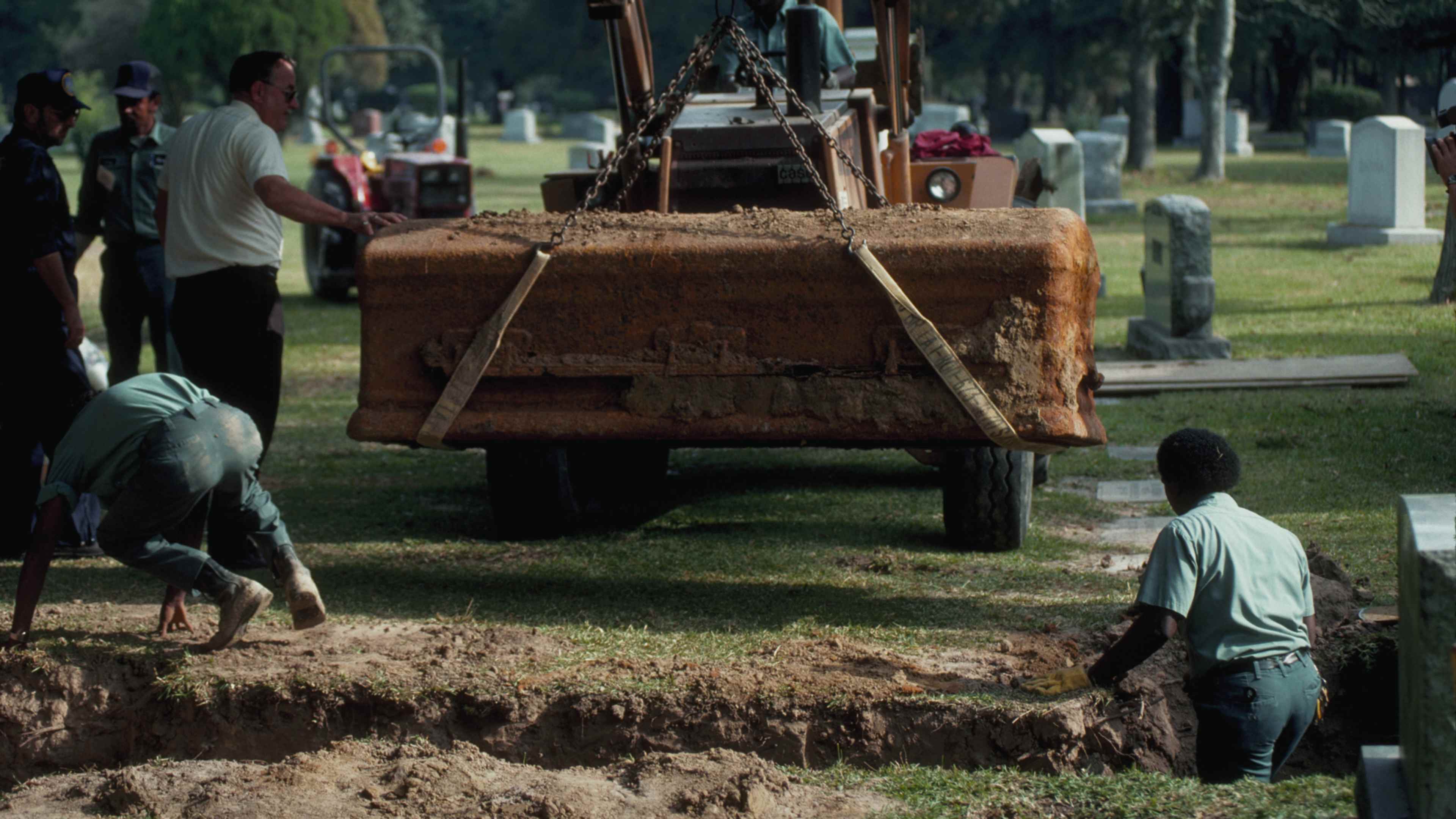 A construction site in a grassy area, with a large excavator and several workers in the foreground, and a cemetery or graveyard visible in the background.