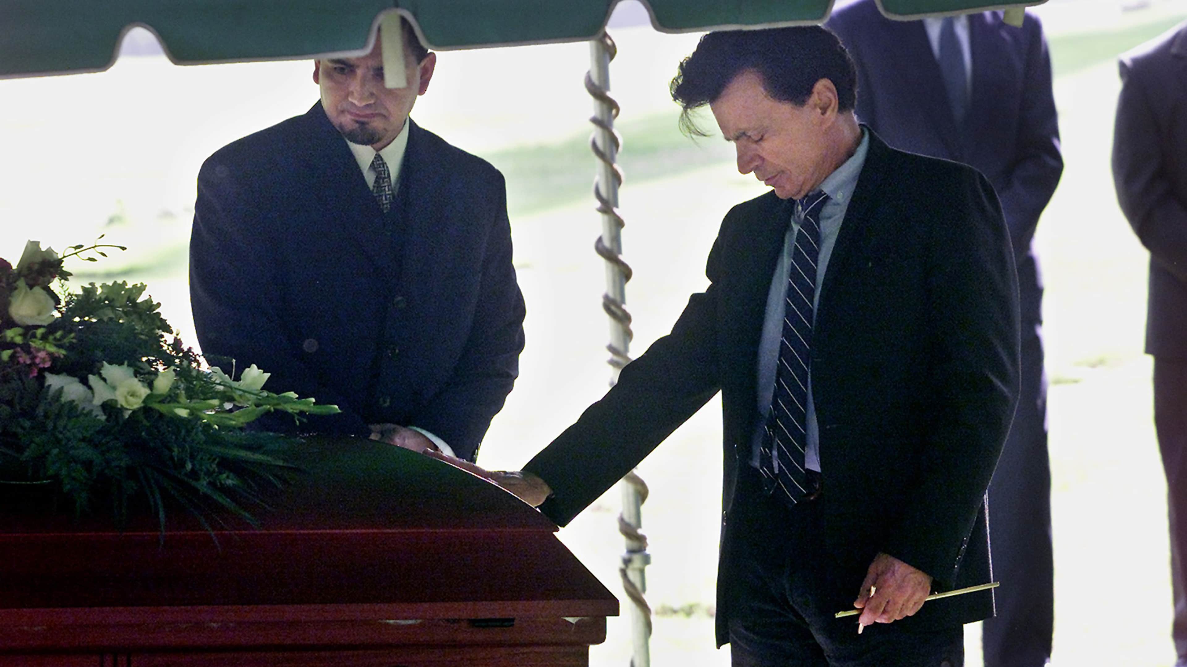 Two men in dark suits standing next to a casket, with a lush, green background visible behind them.