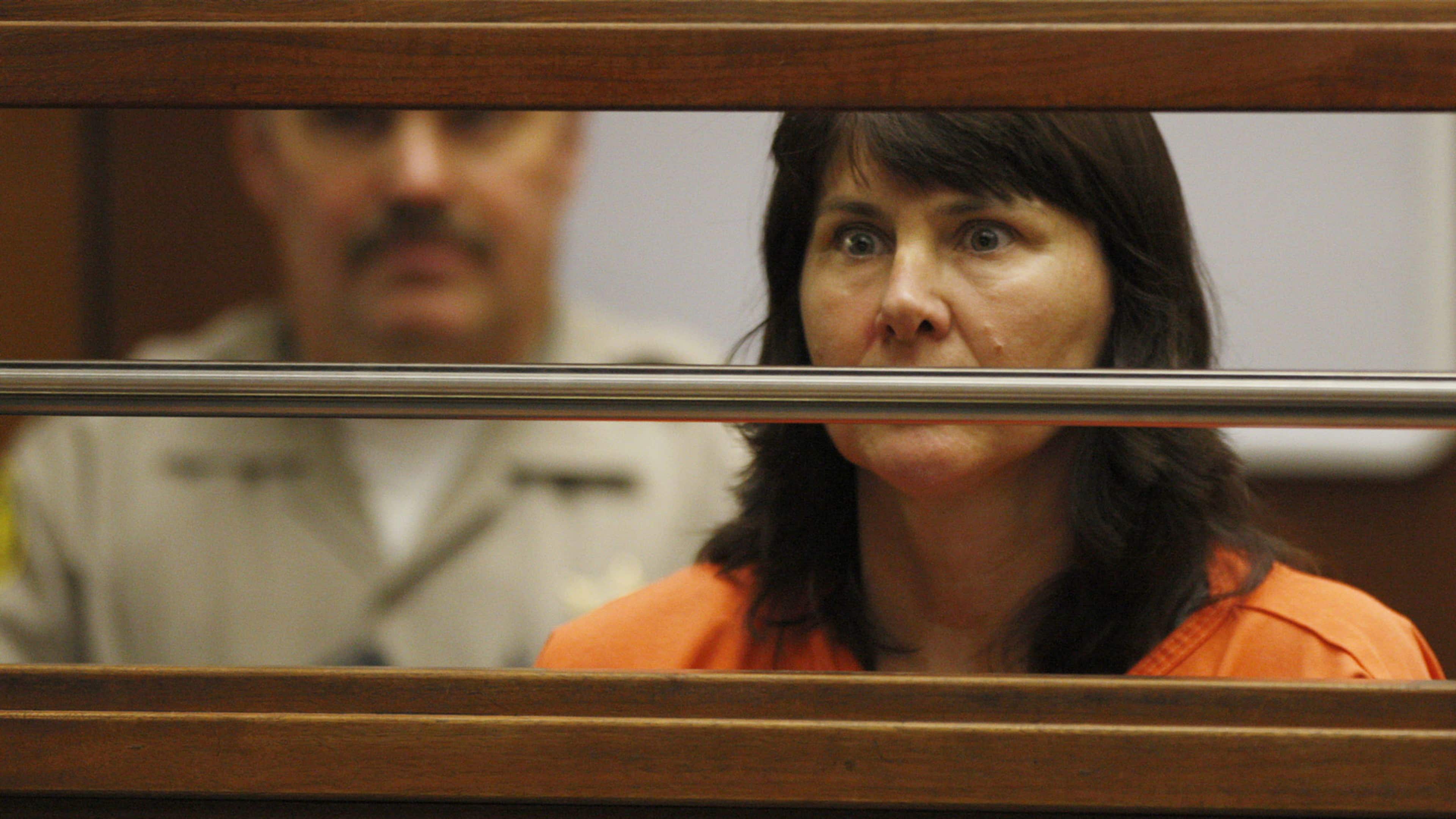 A woman in an orange jumpsuit appears to be in a courtroom setting, with a wooden bench and other individuals visible in the background.