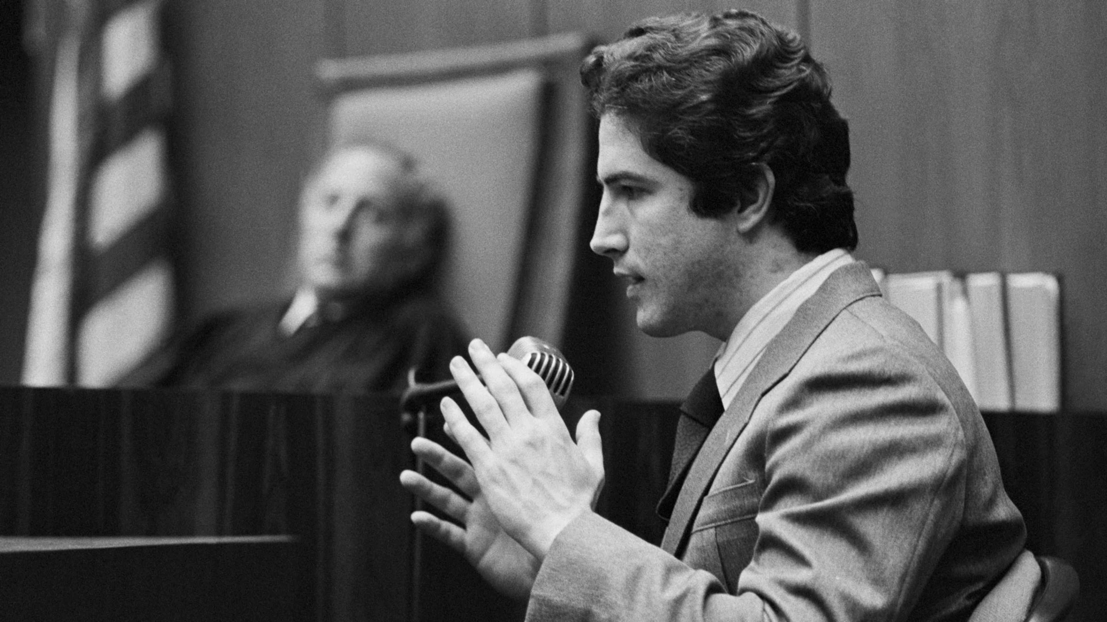 A man in a suit stands in a courtroom, his hands clasped in front of him, with a portrait of a person visible in the background.