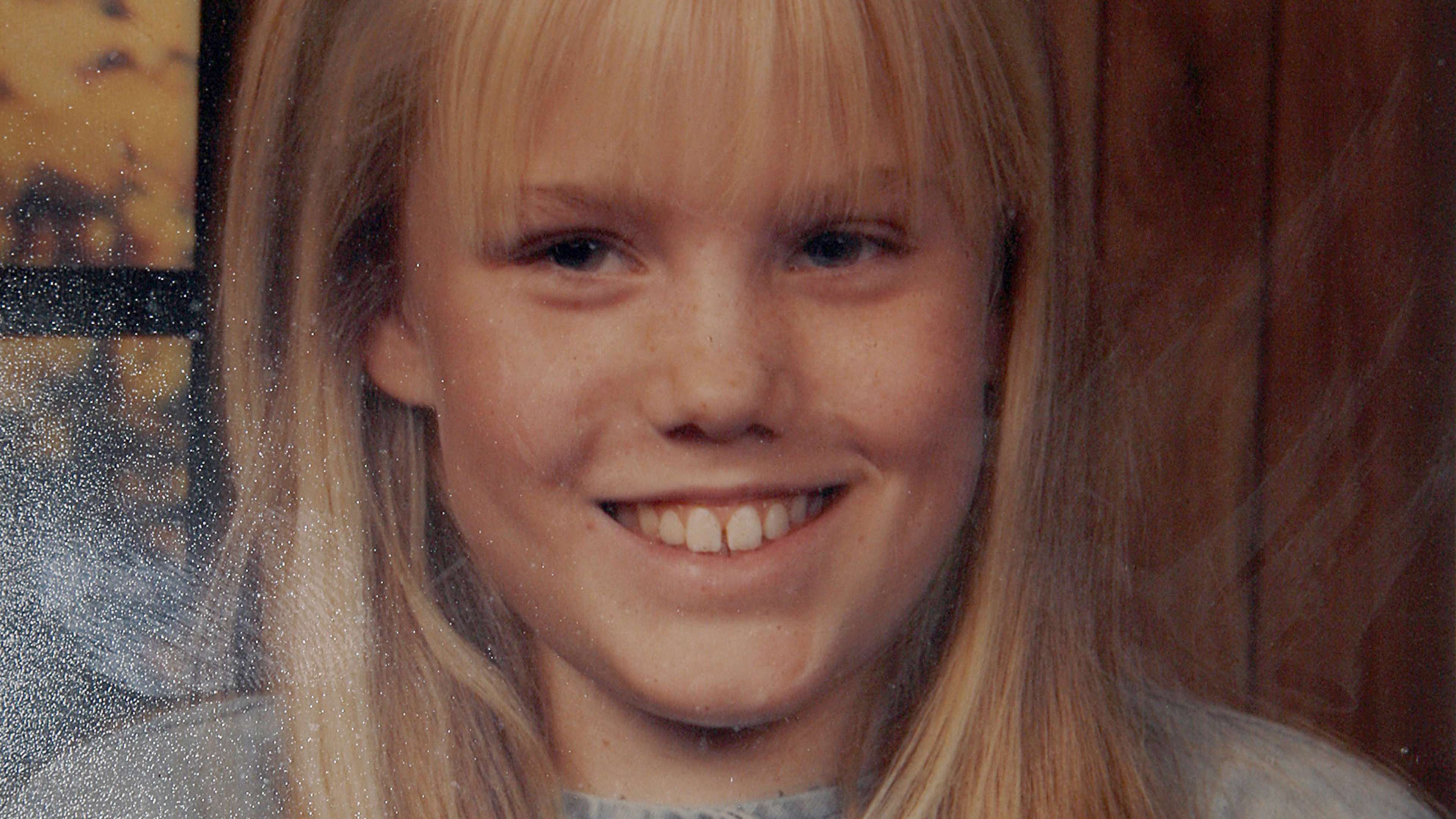 A smiling young woman with long blonde hair appears in the foreground, with a blurred background of what seems to be a window or glass surface.