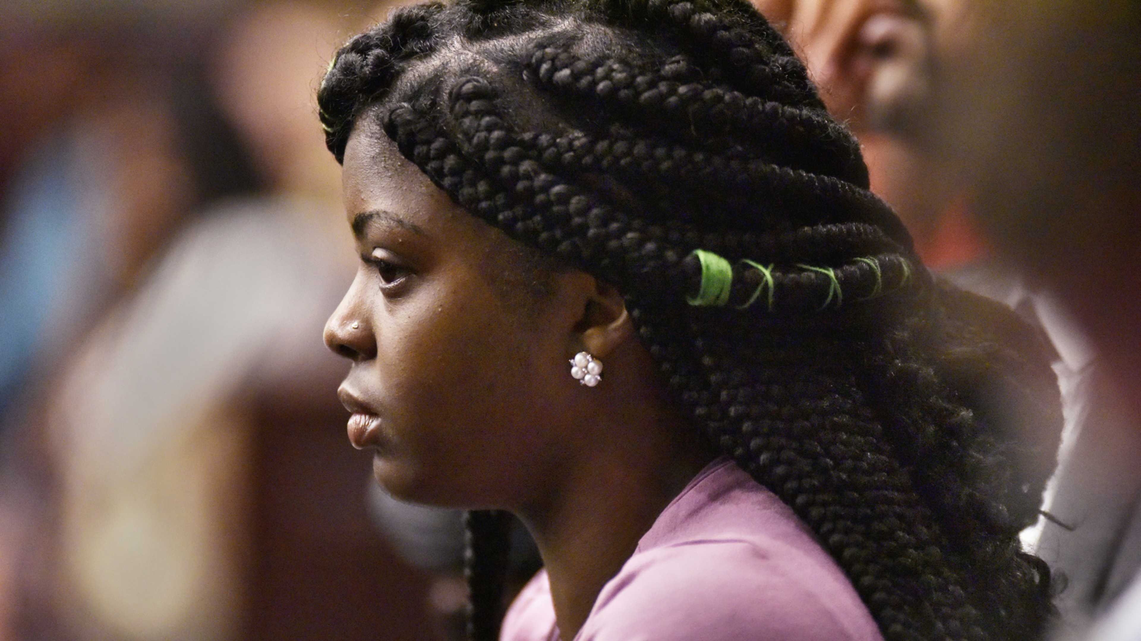 A young woman with intricate braided hairstyle, wearing a pink shirt, appears in the foreground against a blurred background.