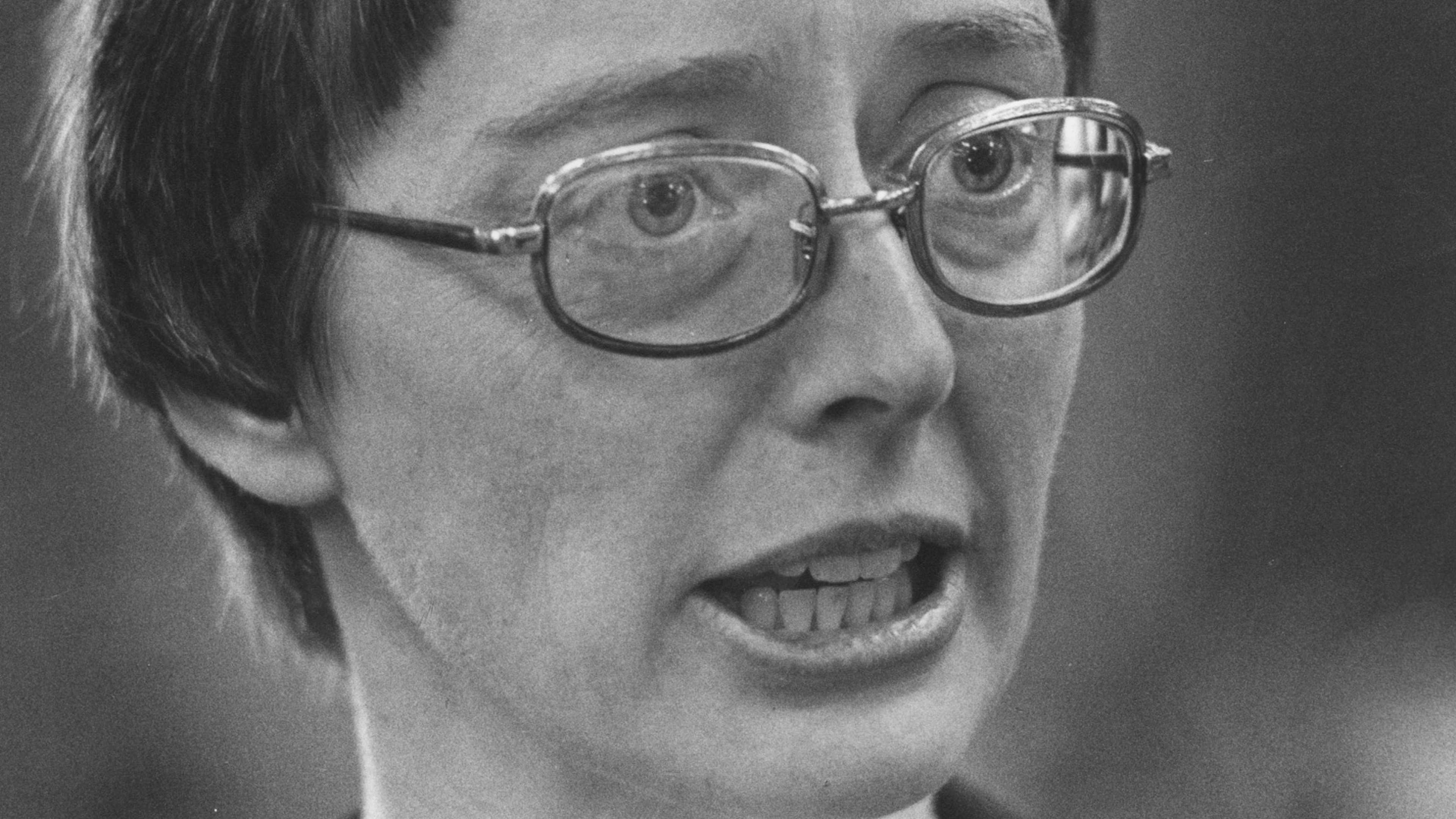 A close-up black and white portrait of a person with glasses, their mouth open in an expression of surprise or excitement.