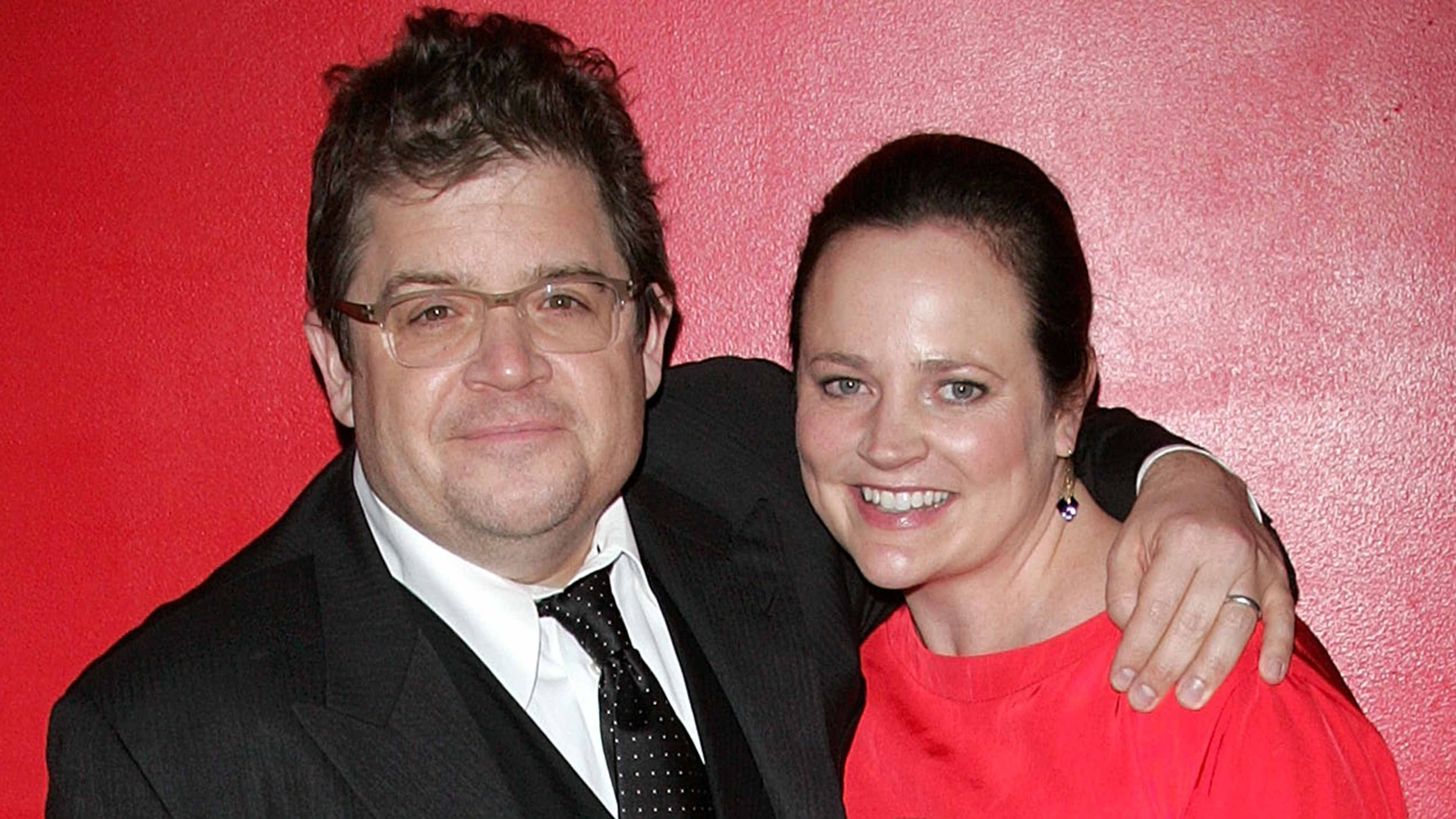The image shows a middle-aged man and woman posing together in front of a vibrant red background, both dressed formally and smiling at the camera.