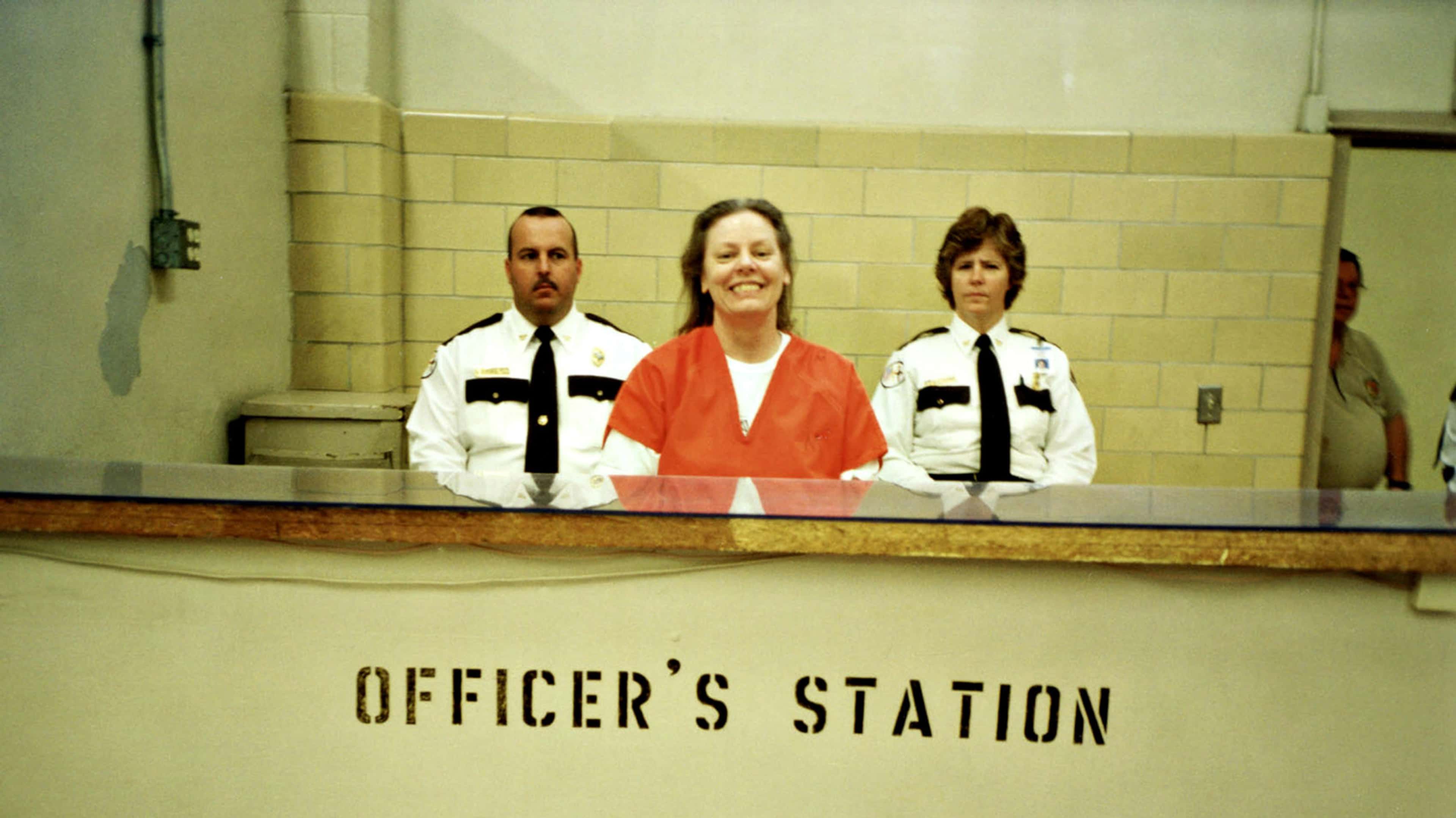 The image shows three individuals, two police officers in uniform and a person in an orange jumpsuit, standing behind a counter labeled "Officer's Station".
