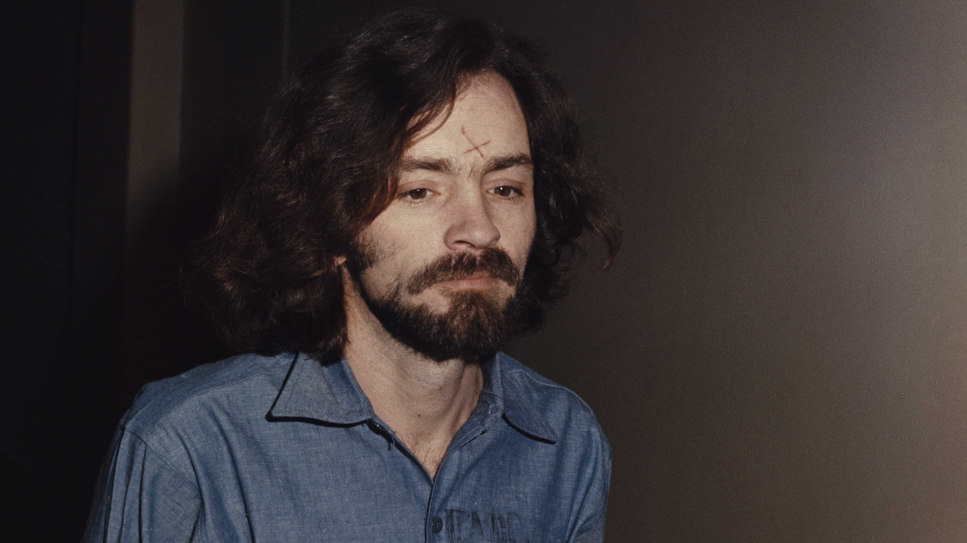 A man with long, dark hair and a beard is shown in a dimly lit setting, his expression appearing pensive or contemplative.