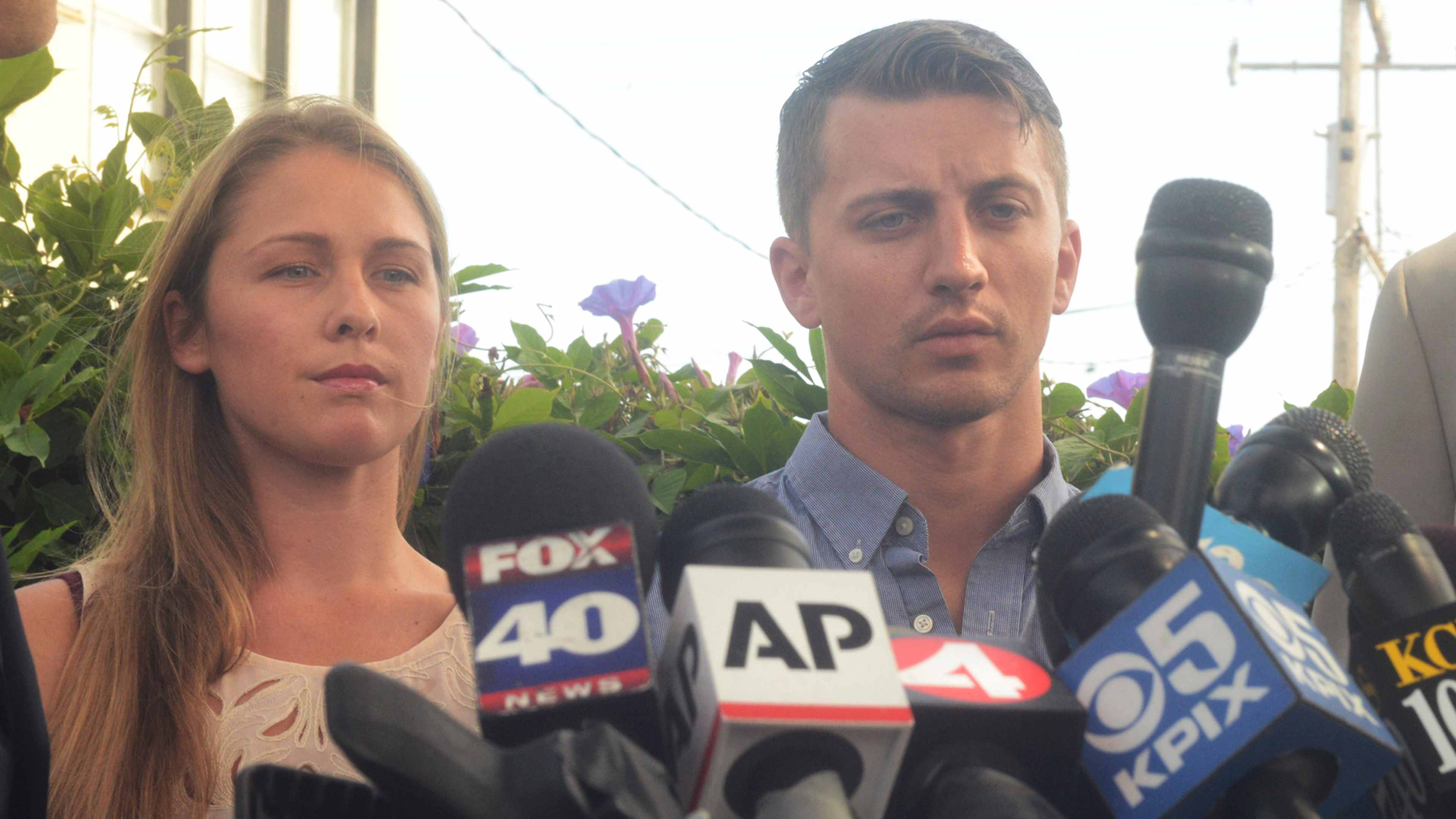 The image shows two people, a woman and a man, standing in front of various news microphones and cameras, suggesting they are being interviewed or giving a statement.