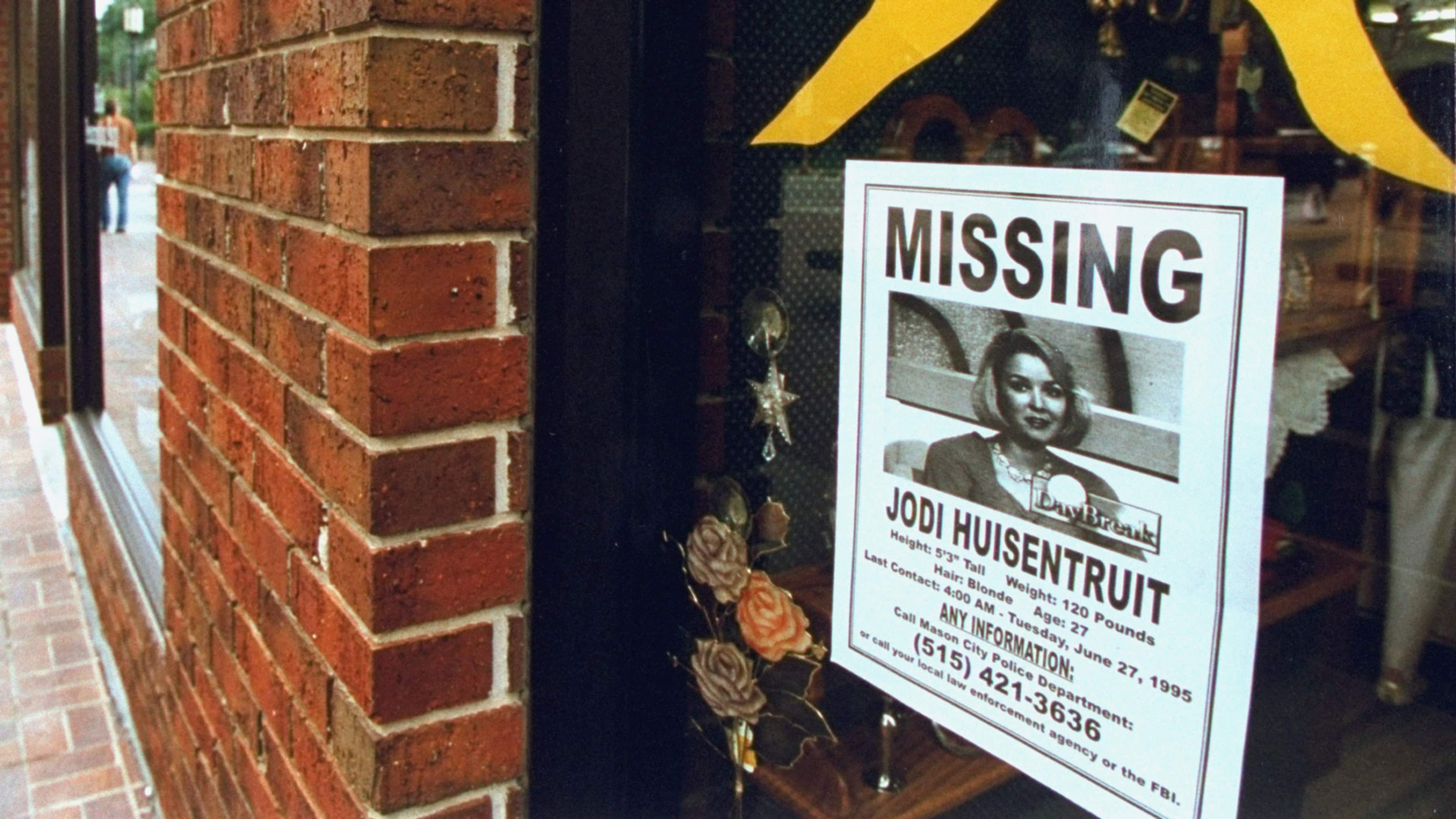 A missing person poster is displayed on a brick wall, featuring a photograph of a woman and contact information for reporting any information about her whereabouts.