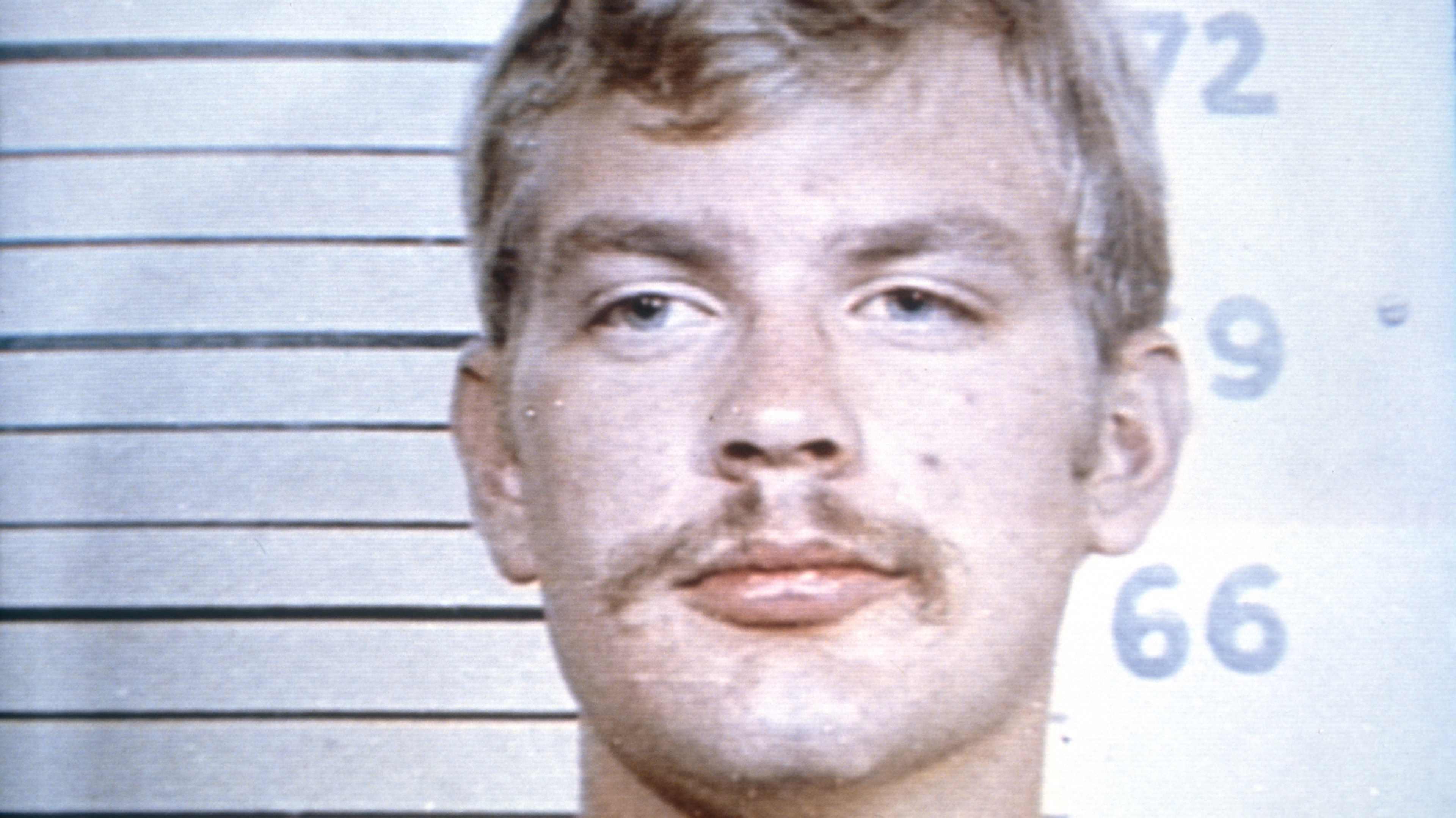 The image shows a close-up portrait of a man with a serious expression, standing in front of a background with numerical markings, suggesting a police lineup or mugshot setting.
