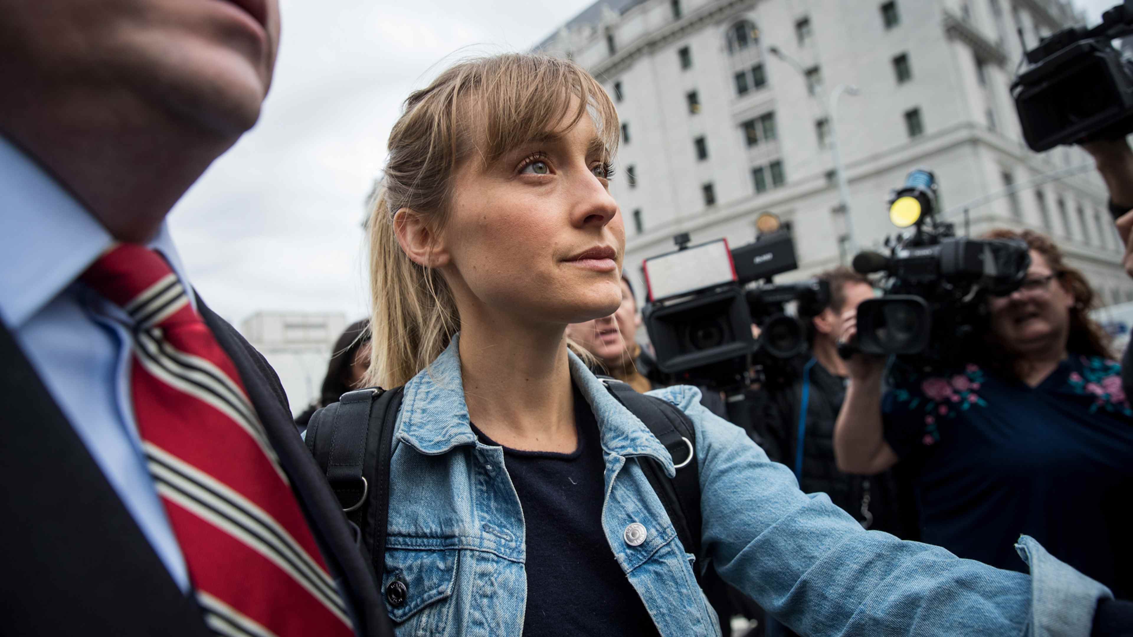A woman in a denim jacket stands in the foreground, surrounded by photographers and camera equipment in an urban setting with tall buildings in the background.