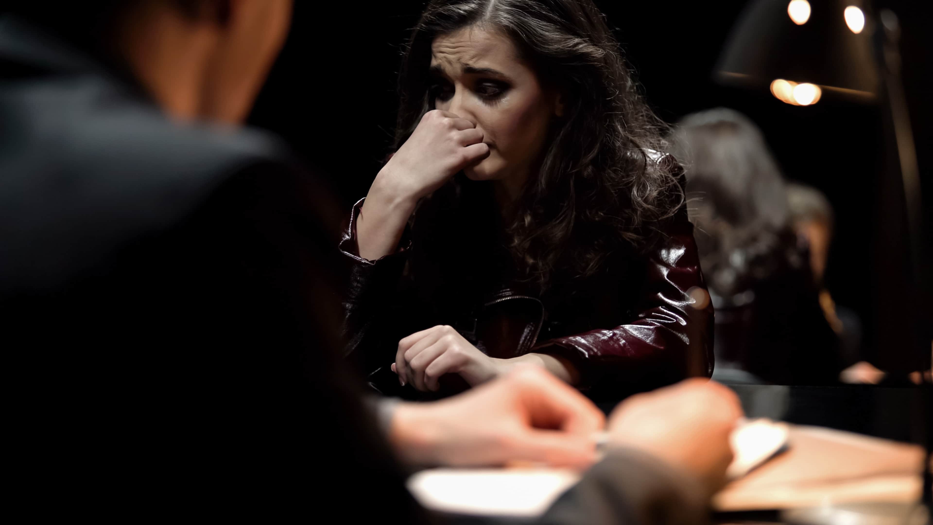 A woman with long, dark hair sits in a dimly lit room, her face partially obscured by her hand as she appears to be in a pensive or emotional state.