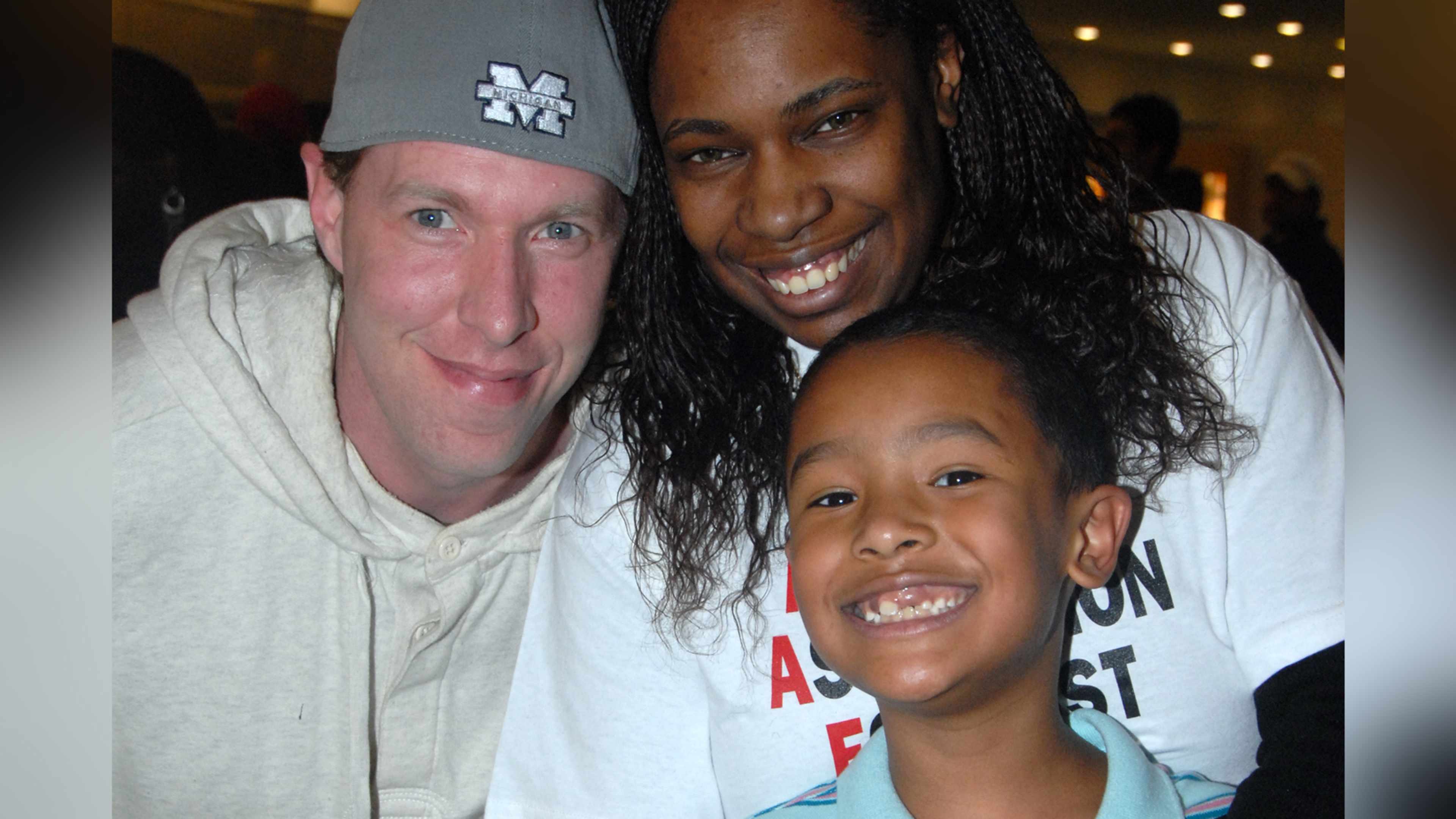 The image shows a smiling group of three people, two adults and one child, posing together in what appears to be a casual setting with some lighting fixtures visible in the background.