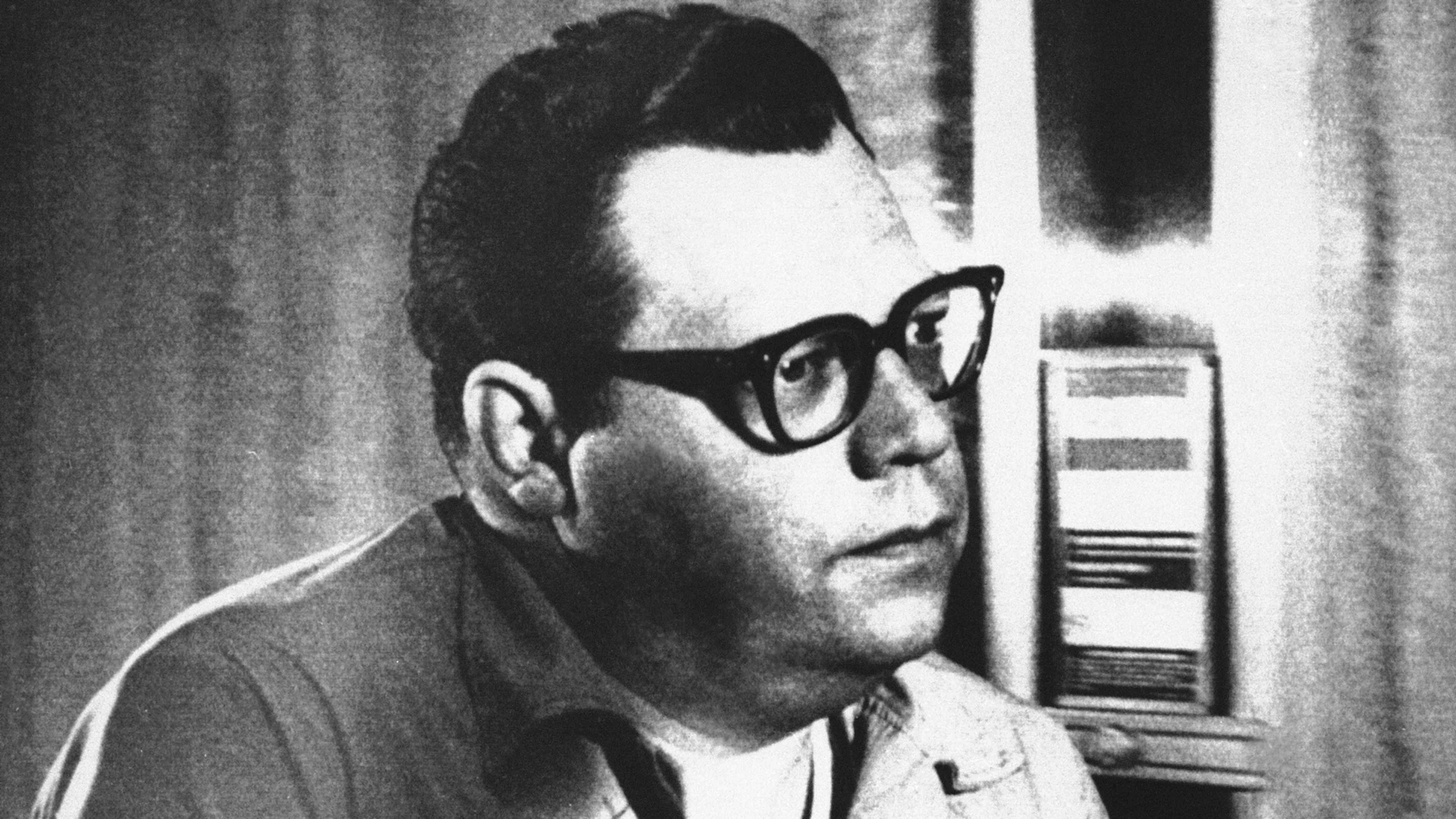 A man with glasses and a serious expression, standing in front of a curtain or wall.