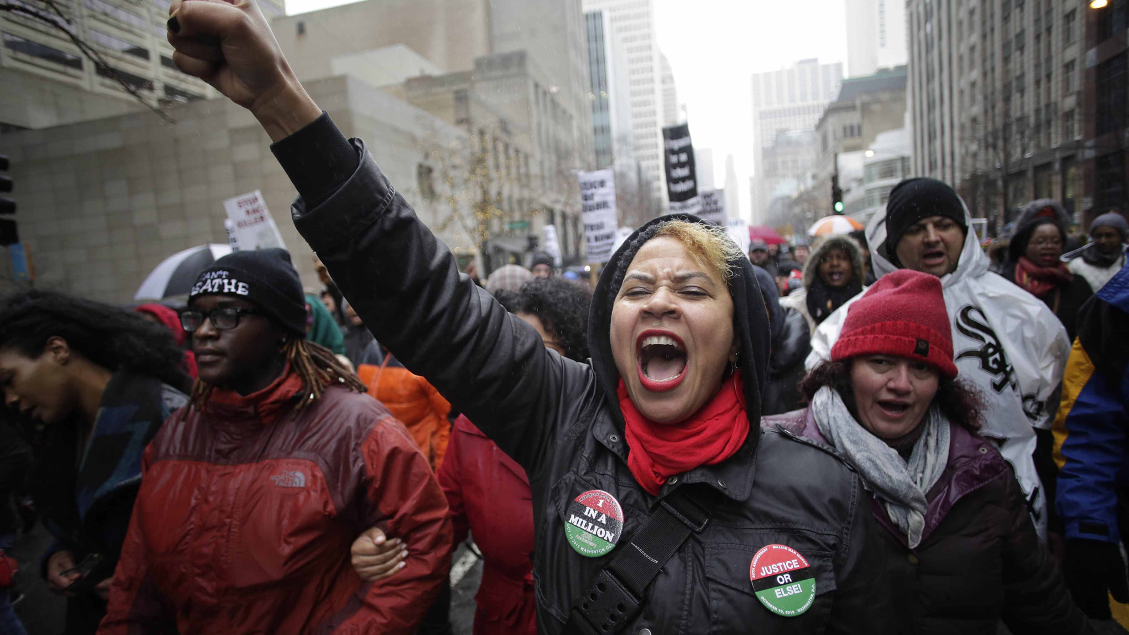 A group of people, some wearing red and black clothing, are gathered in a city street, with their arms raised and expressions of protest or activism.