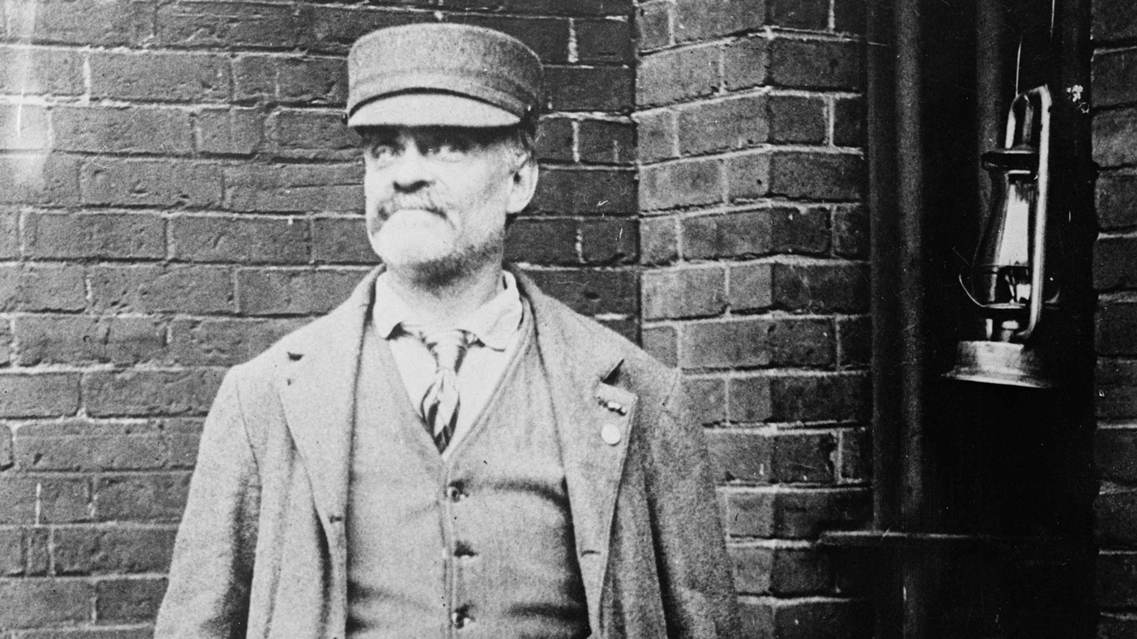 An elderly man with a long beard and a cap stands in front of a brick wall, his expression somber and pensive.